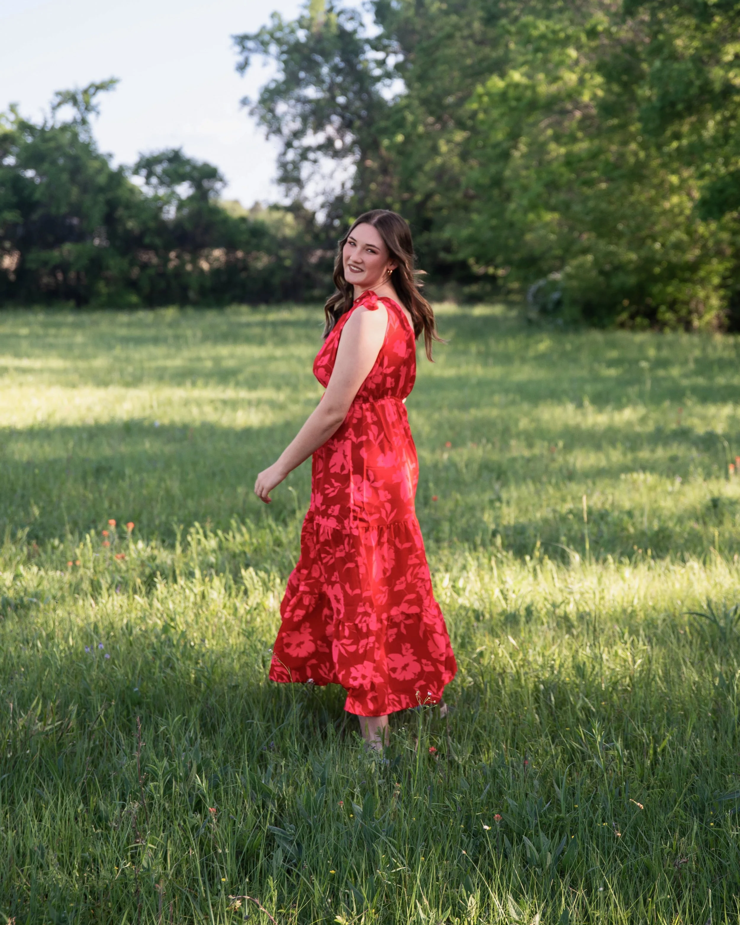 A young woman in a red floral dress standing in a grassy field with trees in the background.