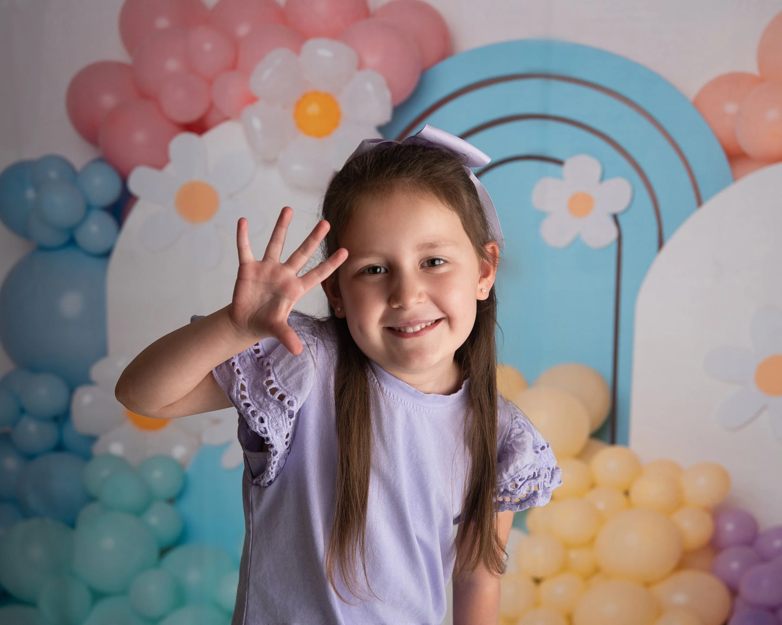 A young girl with long brown hair, wearing a lavender top with lace details on the sleeves, smiling and waving at the camera. She has a purple bow in her hair. The background features colorful balloons and paper decorations with a floral and rainbow 
