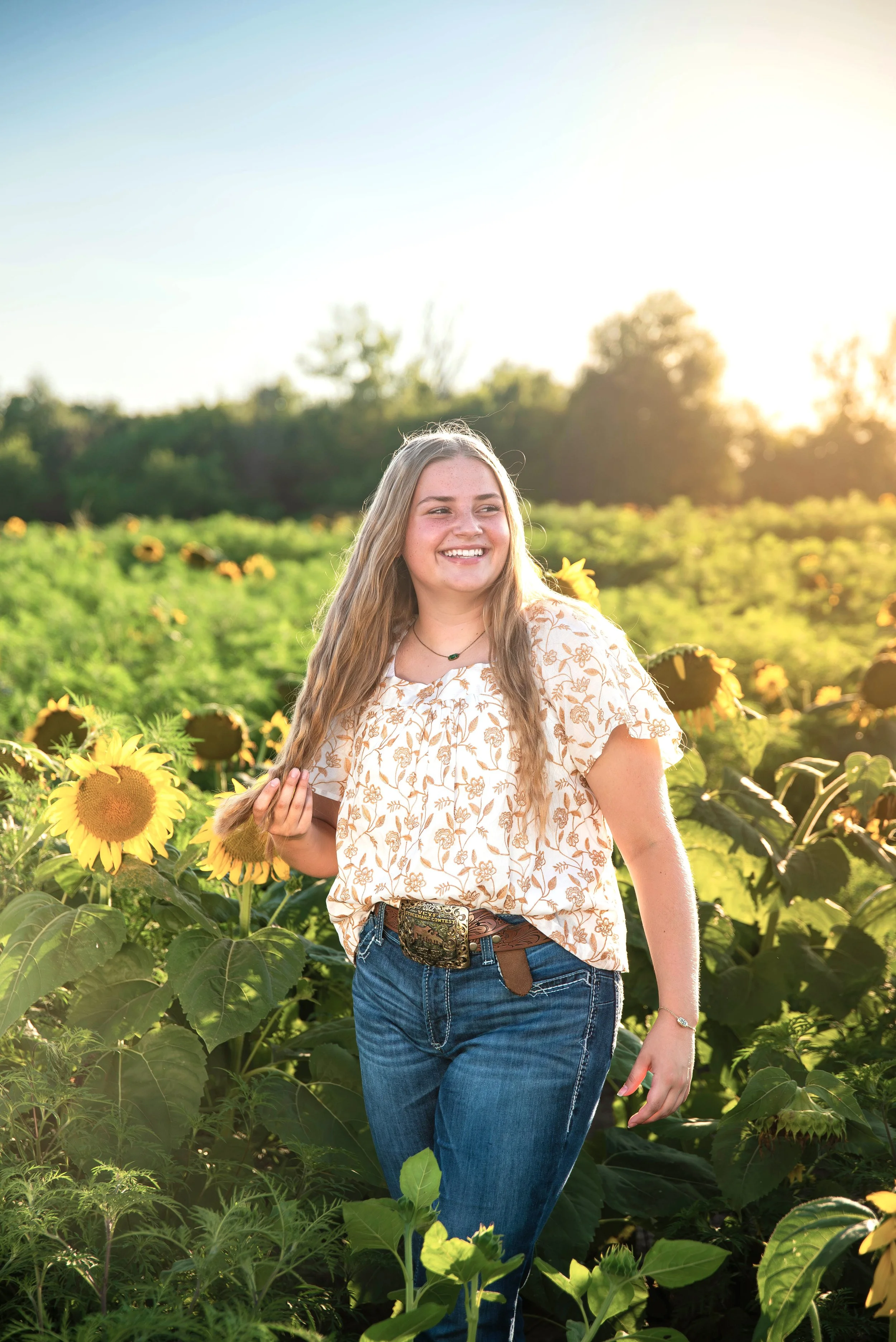 A young woman with long blonde hair, smiling and holding a sunflower, walking in a sunflower field during sunset.