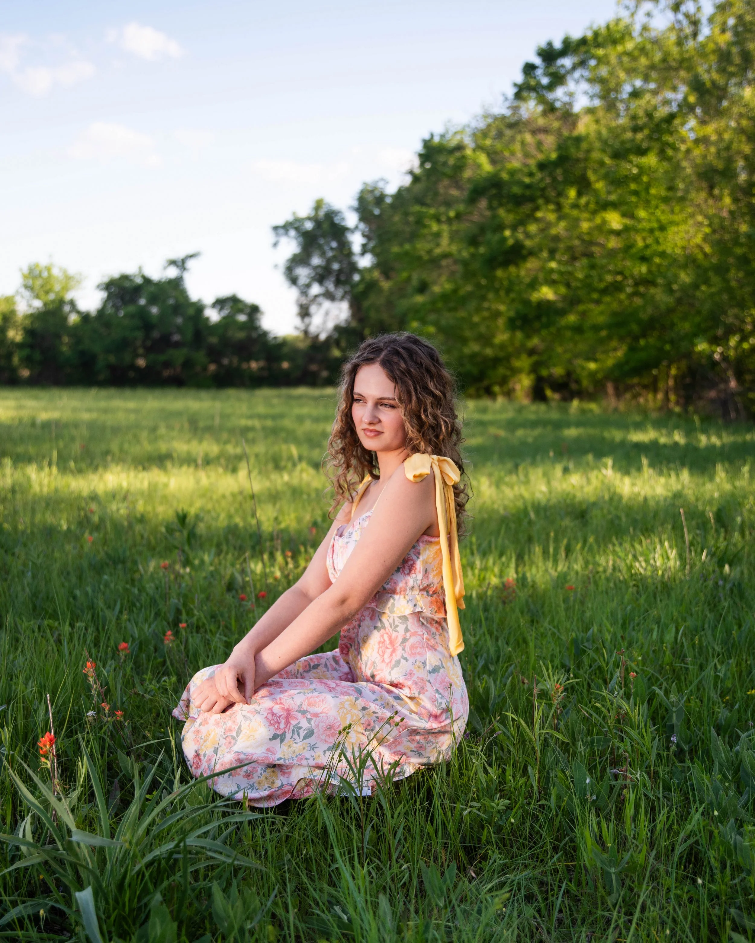 A young woman with curly hair sitting on grass in a field with trees in the background.