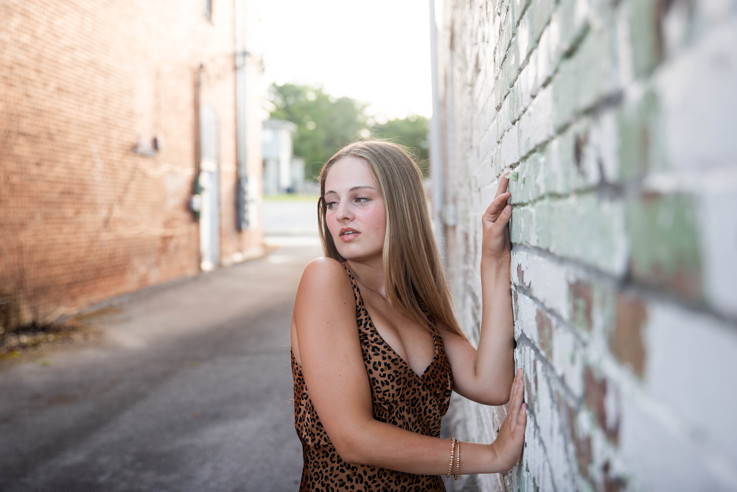 A young woman with long brown hair wearing a leopard print dress leaning against a white brick wall in an alleyway, looking off to the side with a contemplative expression.