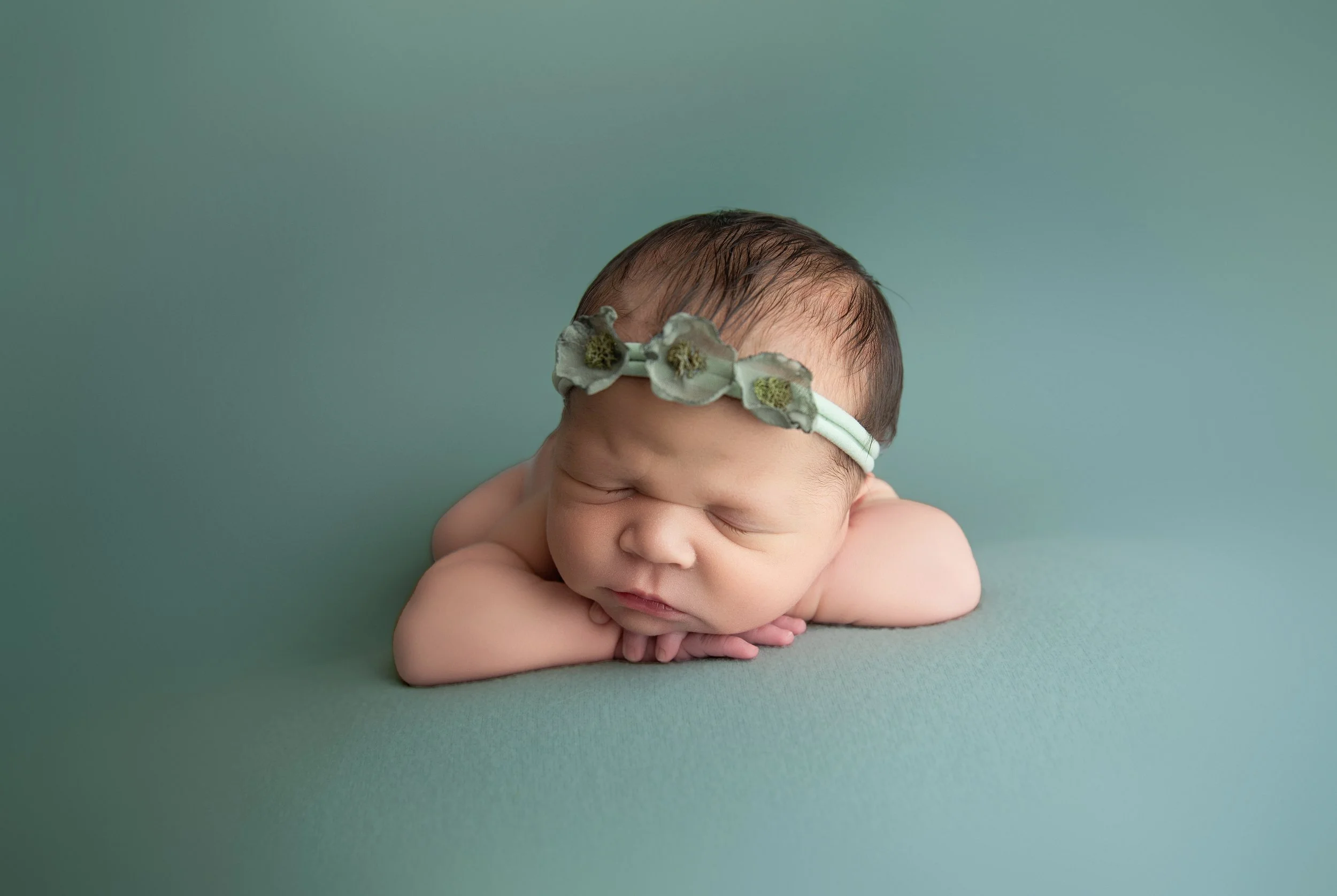 Close-up of a sleeping newborn baby with a floral headband, resting on a soft green surface.