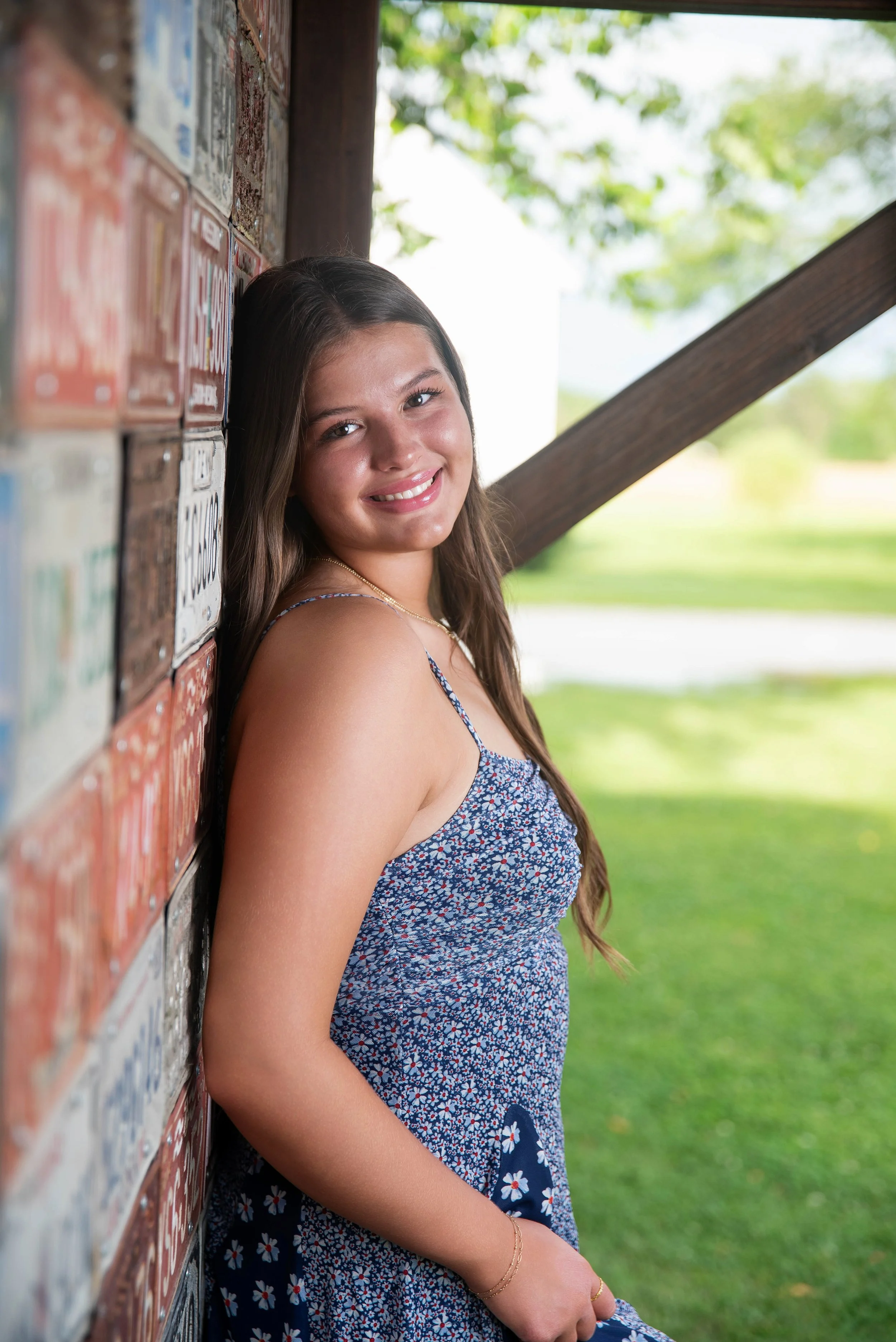 A young woman with long brown hair and a floral dress leaning against a brick wall outdoors smiling at the camera.