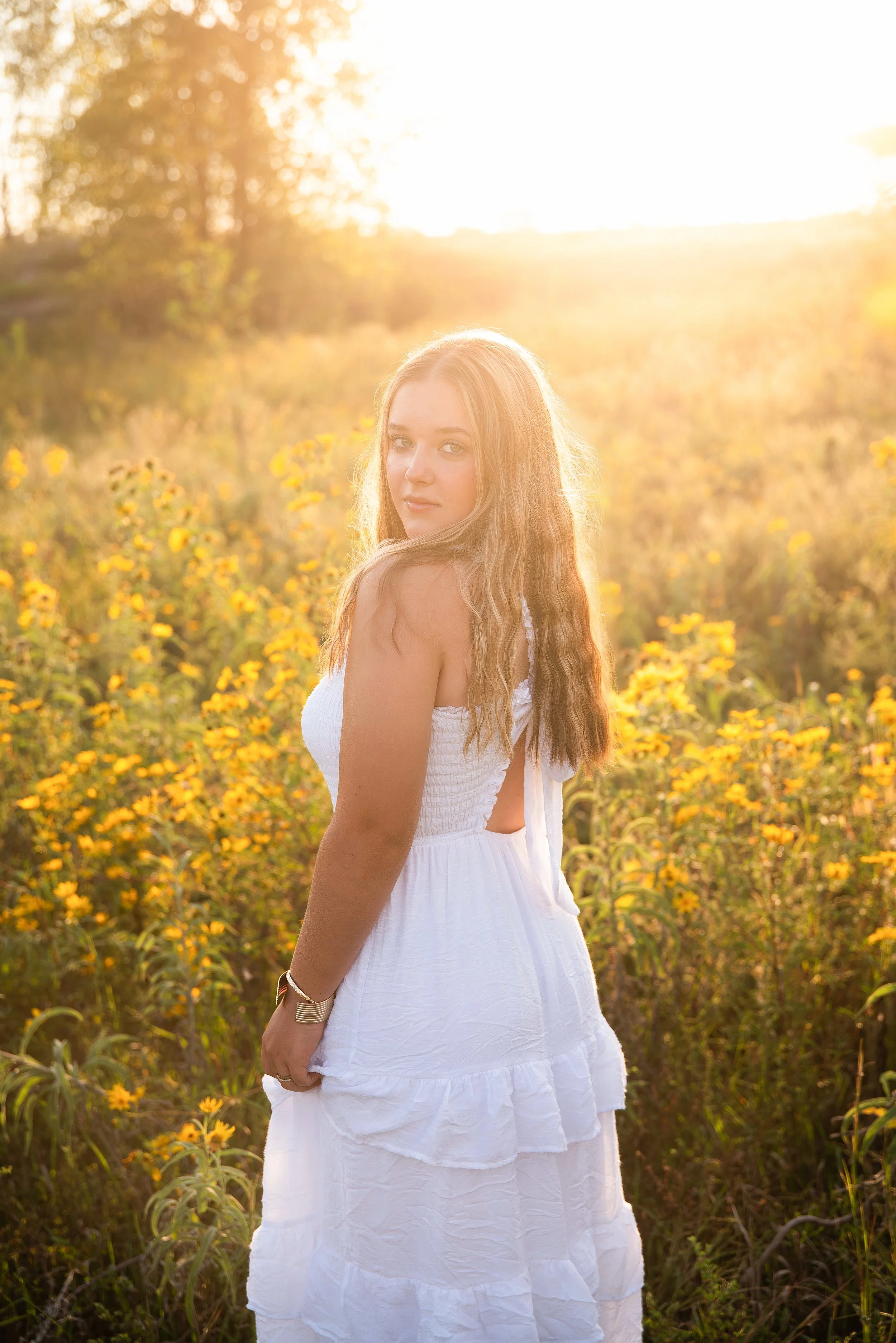 A woman in a white dress standing in a field of yellow flowers during sunset.