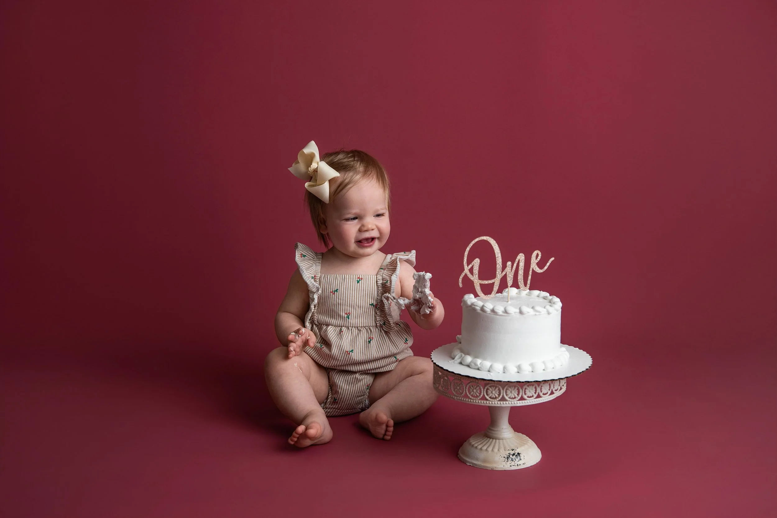 A smiling toddler girl sitting on the floor next to a white birthday cake with a cake topper that spells "One" against a pink background.