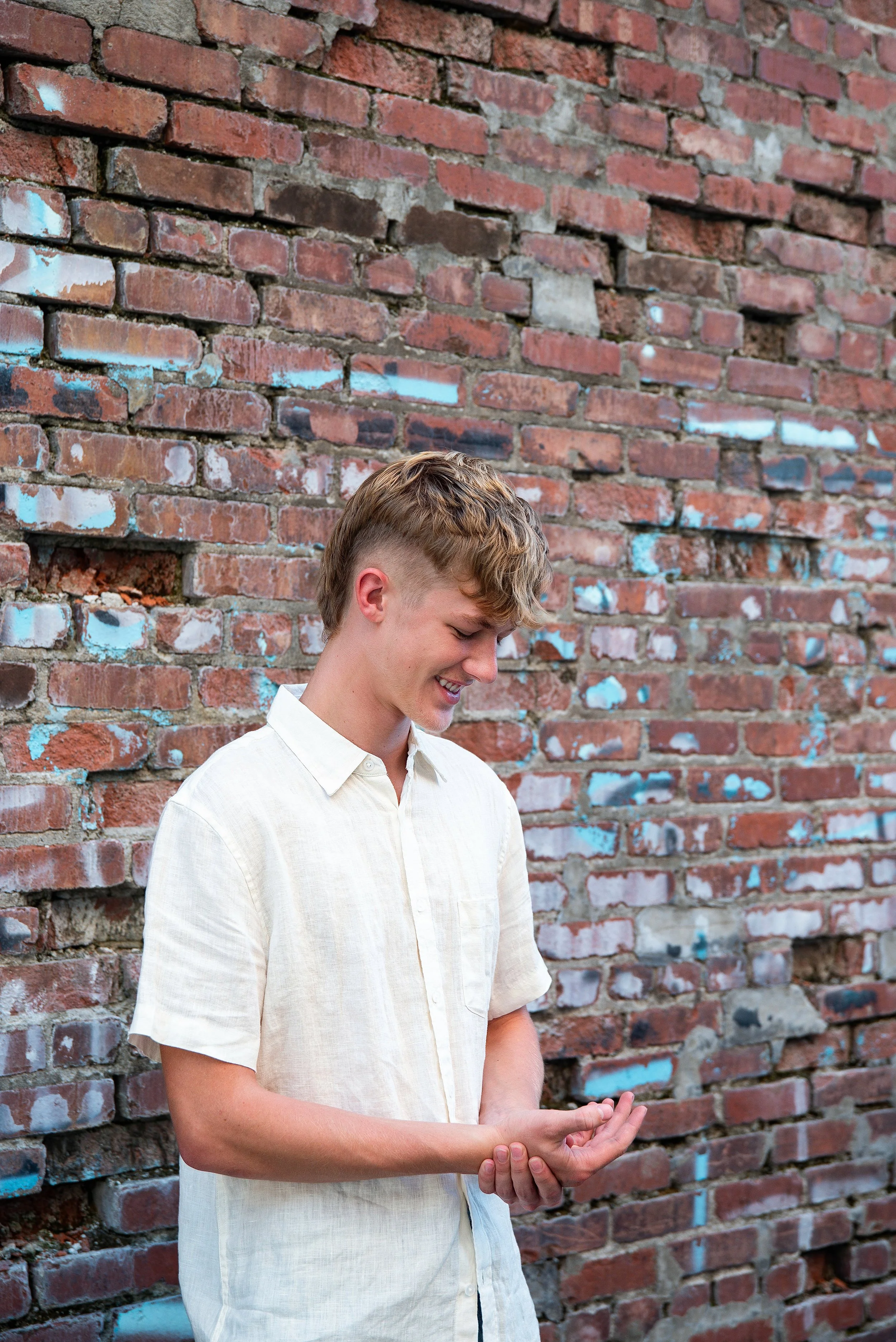 A young man with light brown hair wearing a white short-sleeve shirt, smiling while looking down and holding his hands together in front of a brick wall with some blue graffiti.