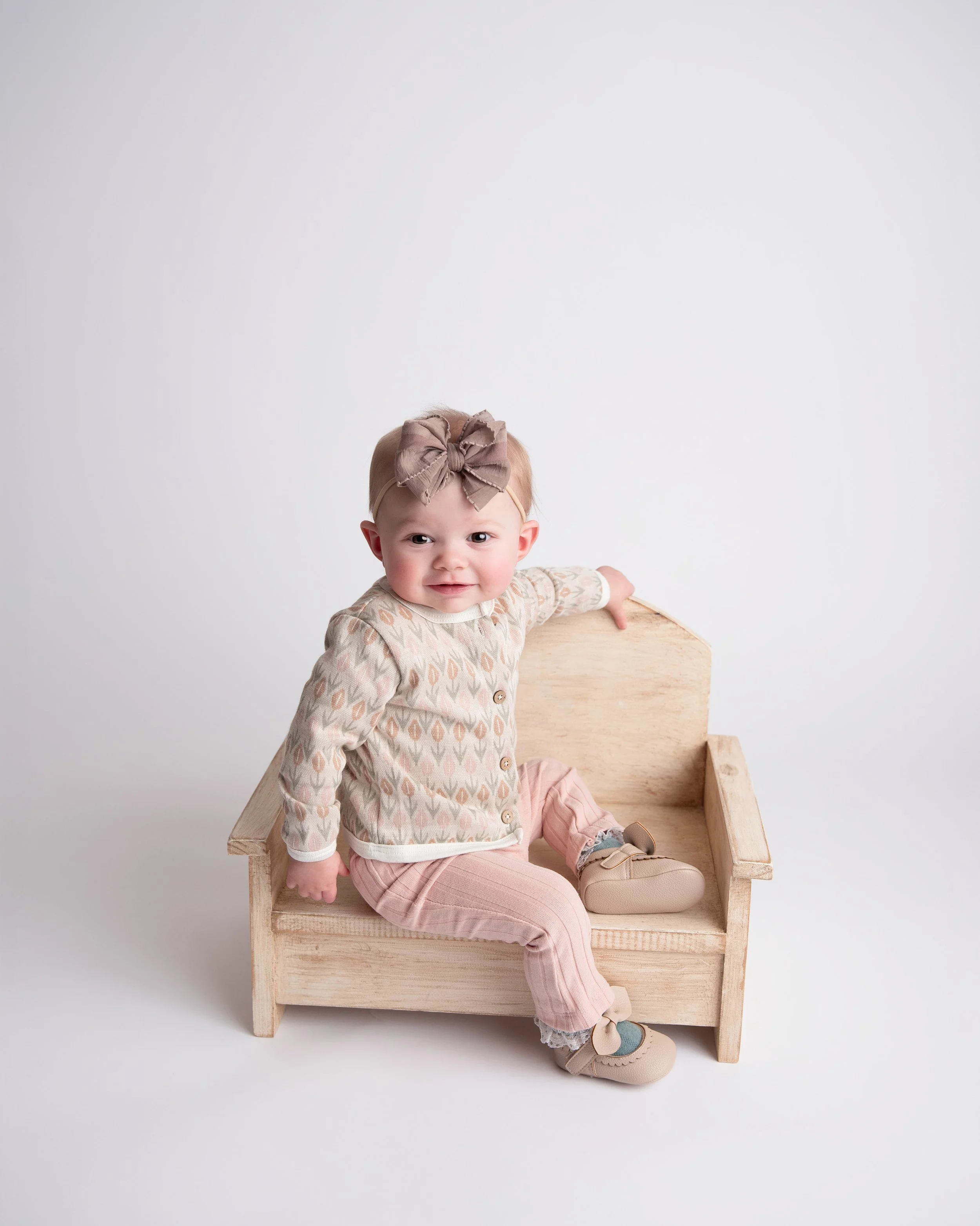 A toddler girl sitting on a small wooden bench, wearing a patterned cardigan, pink pants, and beige shoes with bows, with a large beige bow headband, against a white background.