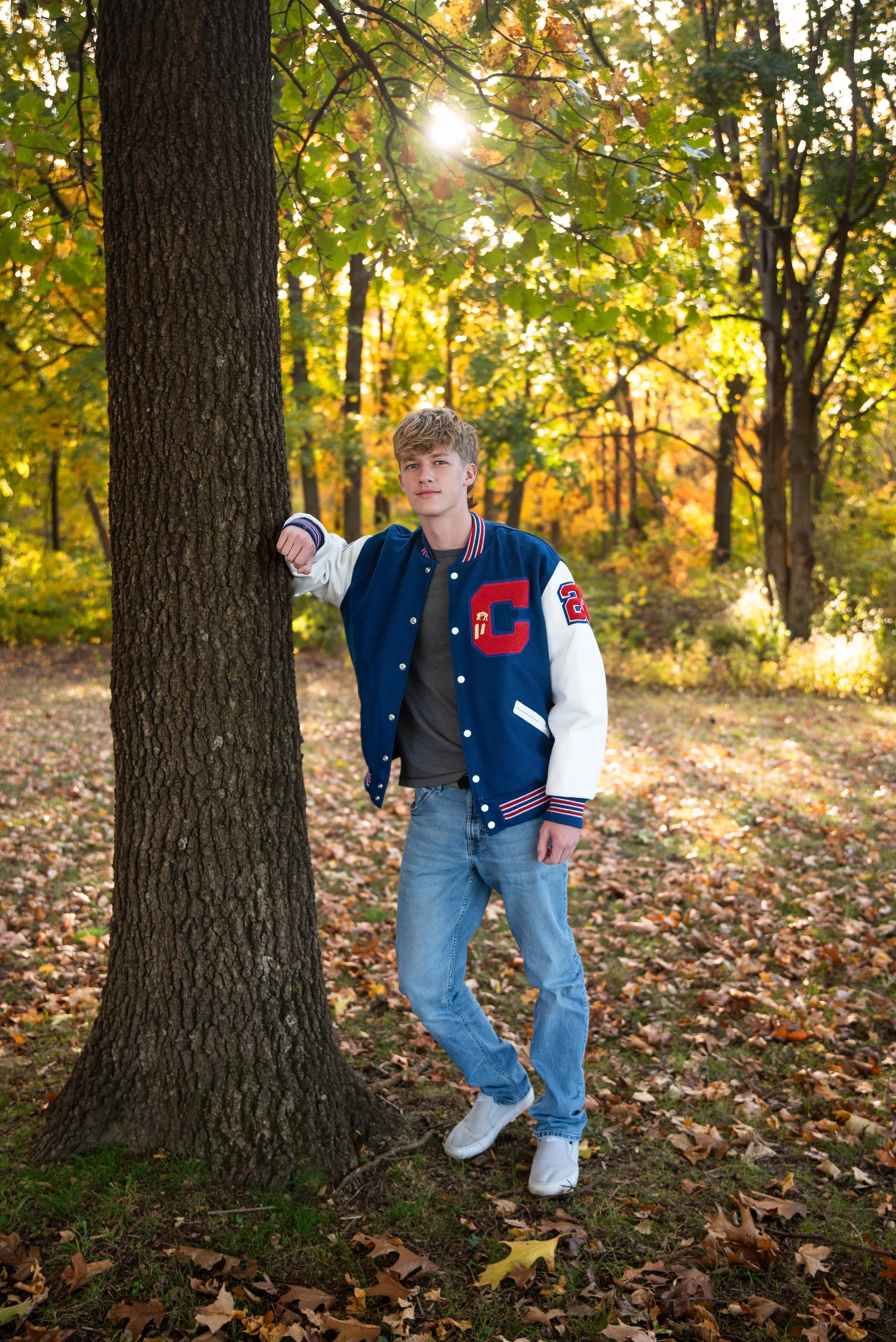 A young man standing next to a tree in a park during autumn. He is leaning with his arm on the tree, wearing a blue and white varsity jacket, grey shirt, blue jeans, and white shoes. The background features colorful fall foliage and sunlight filterin