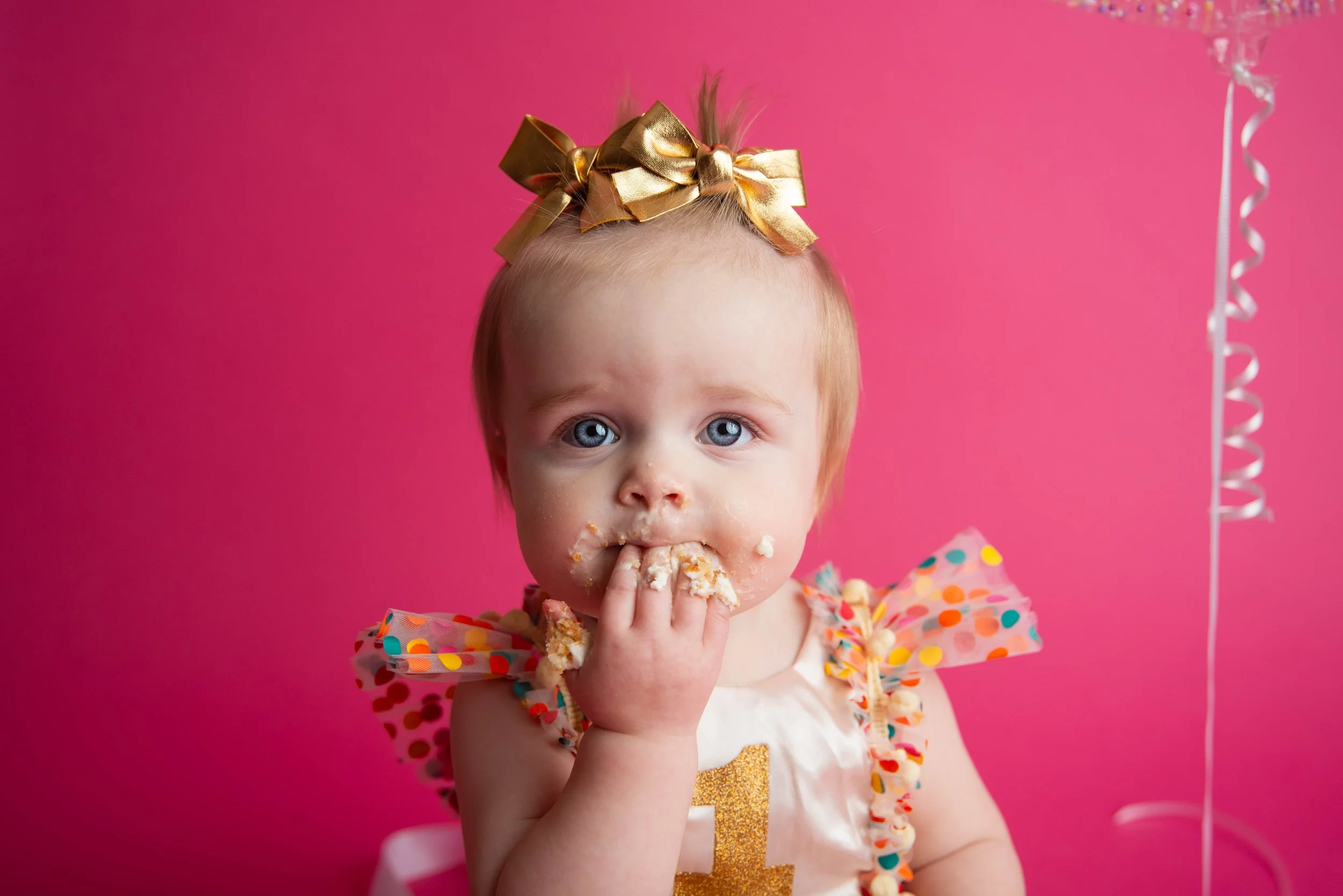 Baby girl with blue eyes and a gold bow in her hair, wearing a cream dress with colorful polka dot ruffles, eating cake against a pink background.