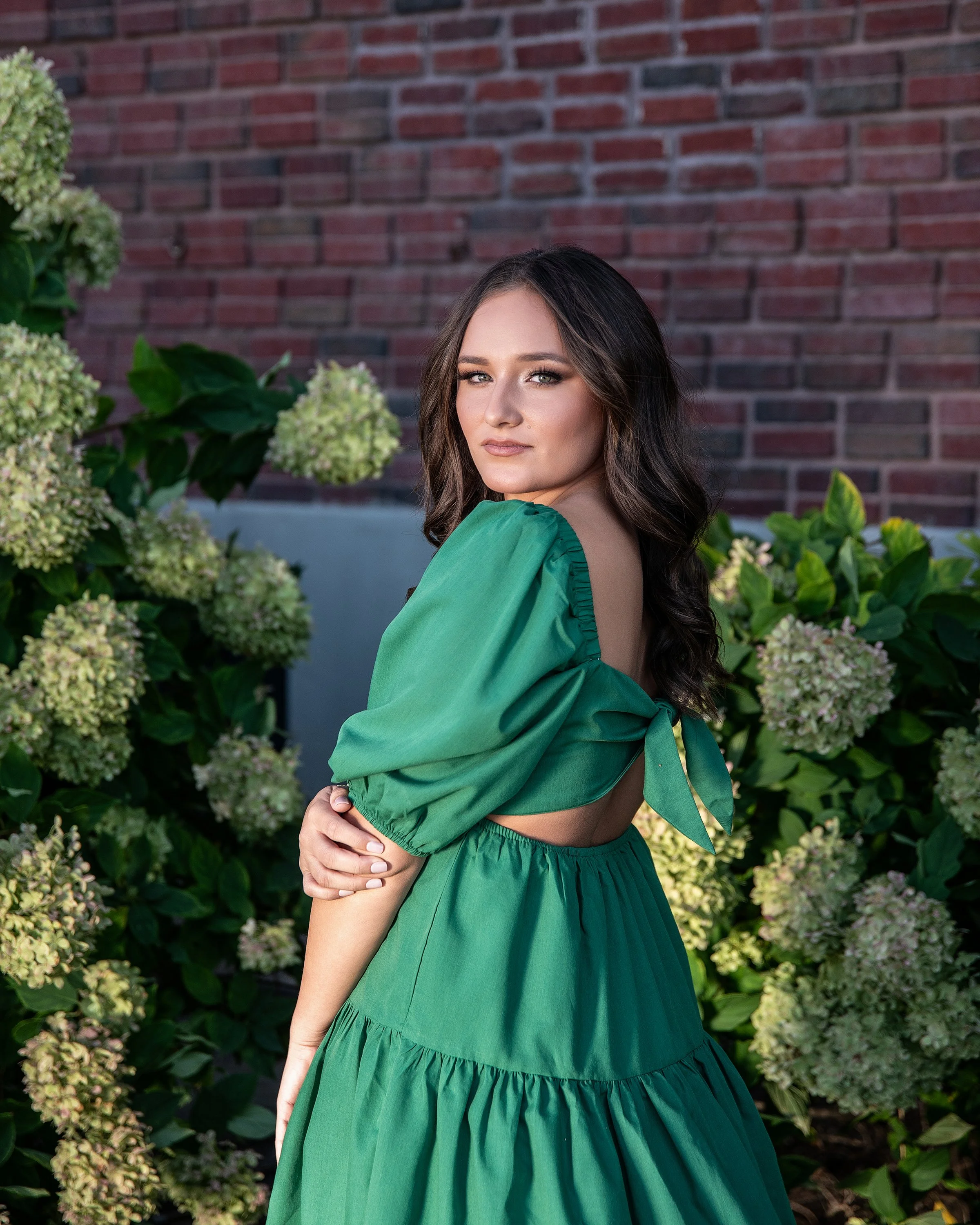 A young woman with dark wavy hair and light makeup posing outdoors in front of green hydrangea bushes and a brick wall, wearing a green dress with puffed sleeves and a tie detail at the back.