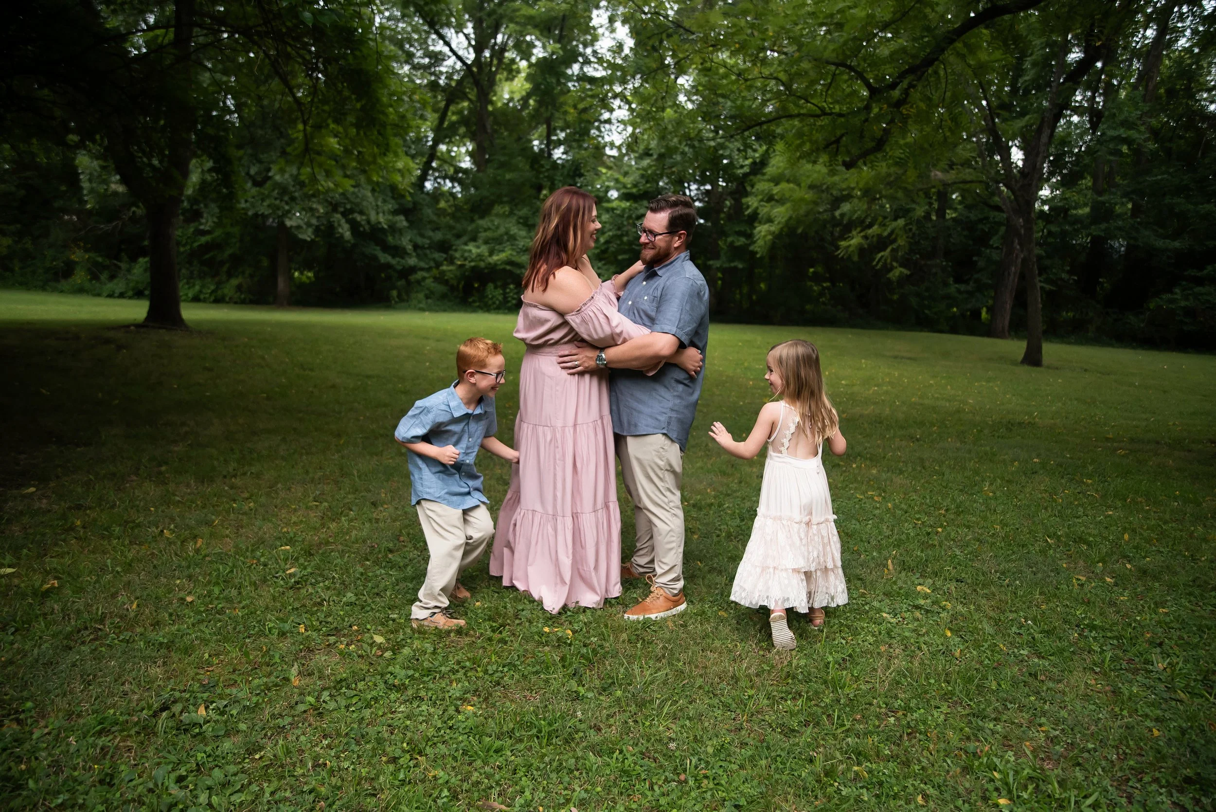 A family of four, including a couple and two children, standing and smiling in a park surrounded by trees and grass.