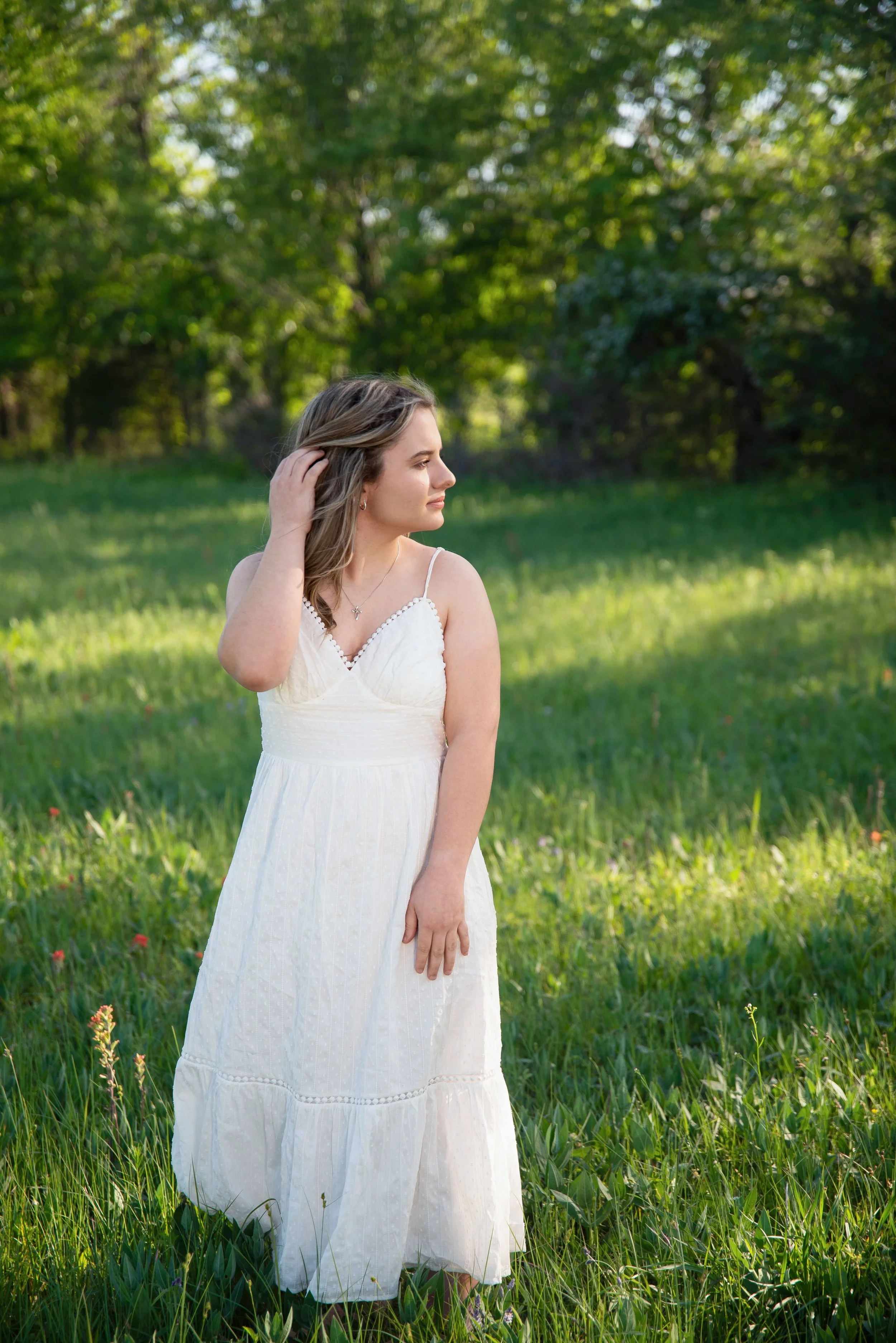 A woman in a white sundress standing in a grassy field with trees in the background. She is looking to the side, touching her hair with one hand.
