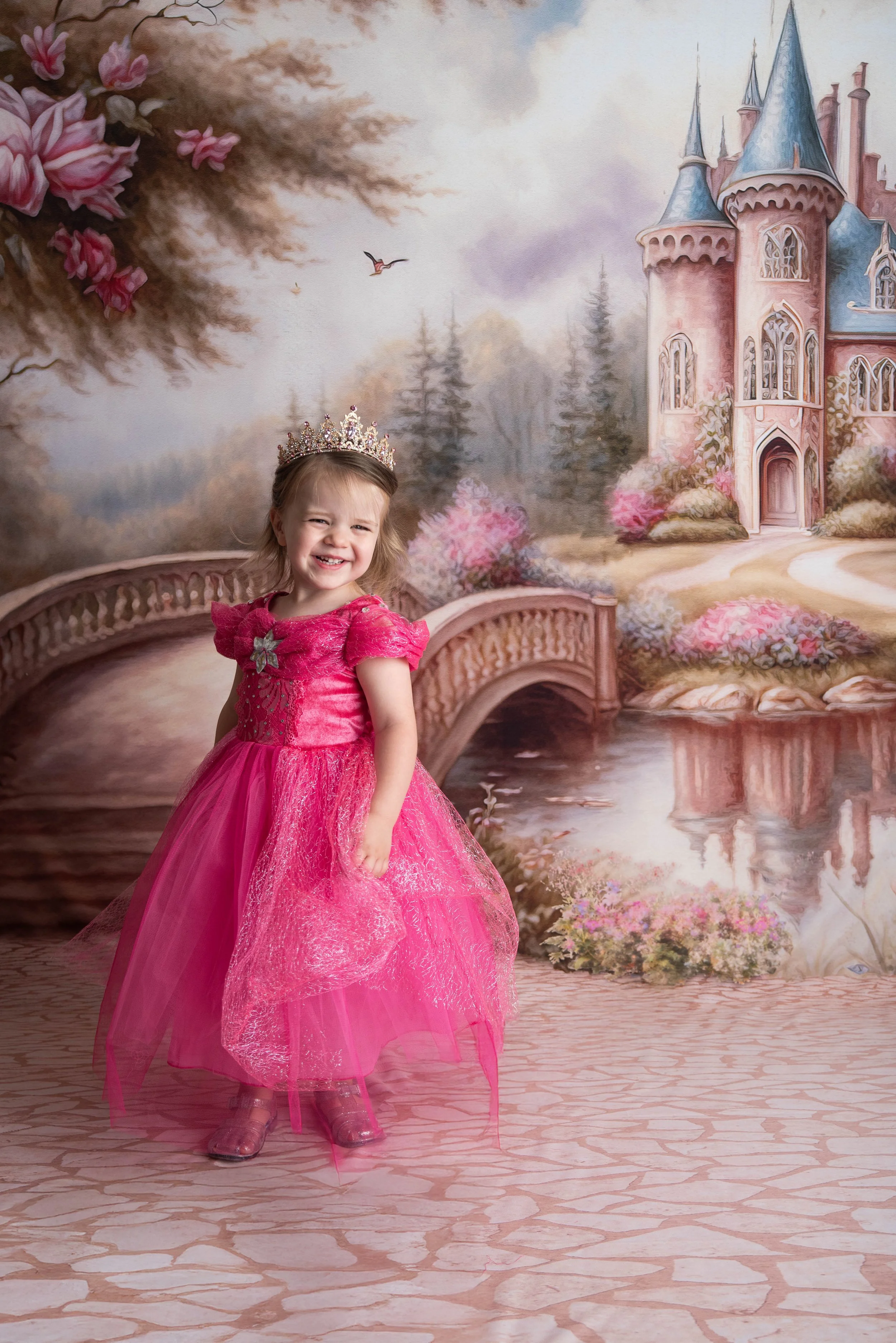 Young girl in a pink princess dress and crown standing in front of a fairy tale castle backdrop.