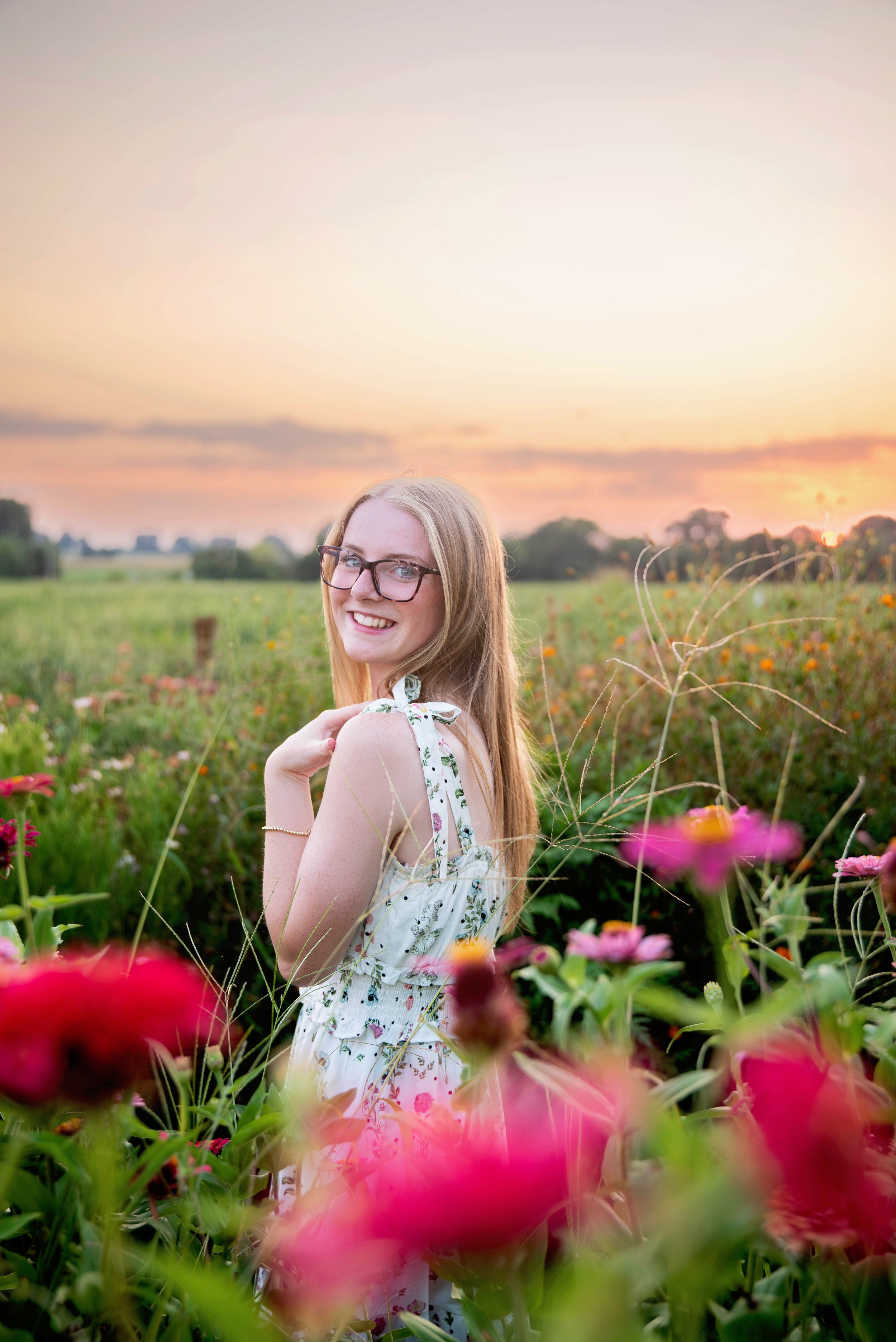 A young woman with glasses and long blonde hair smiling in a field of colorful flowers during sunset.
