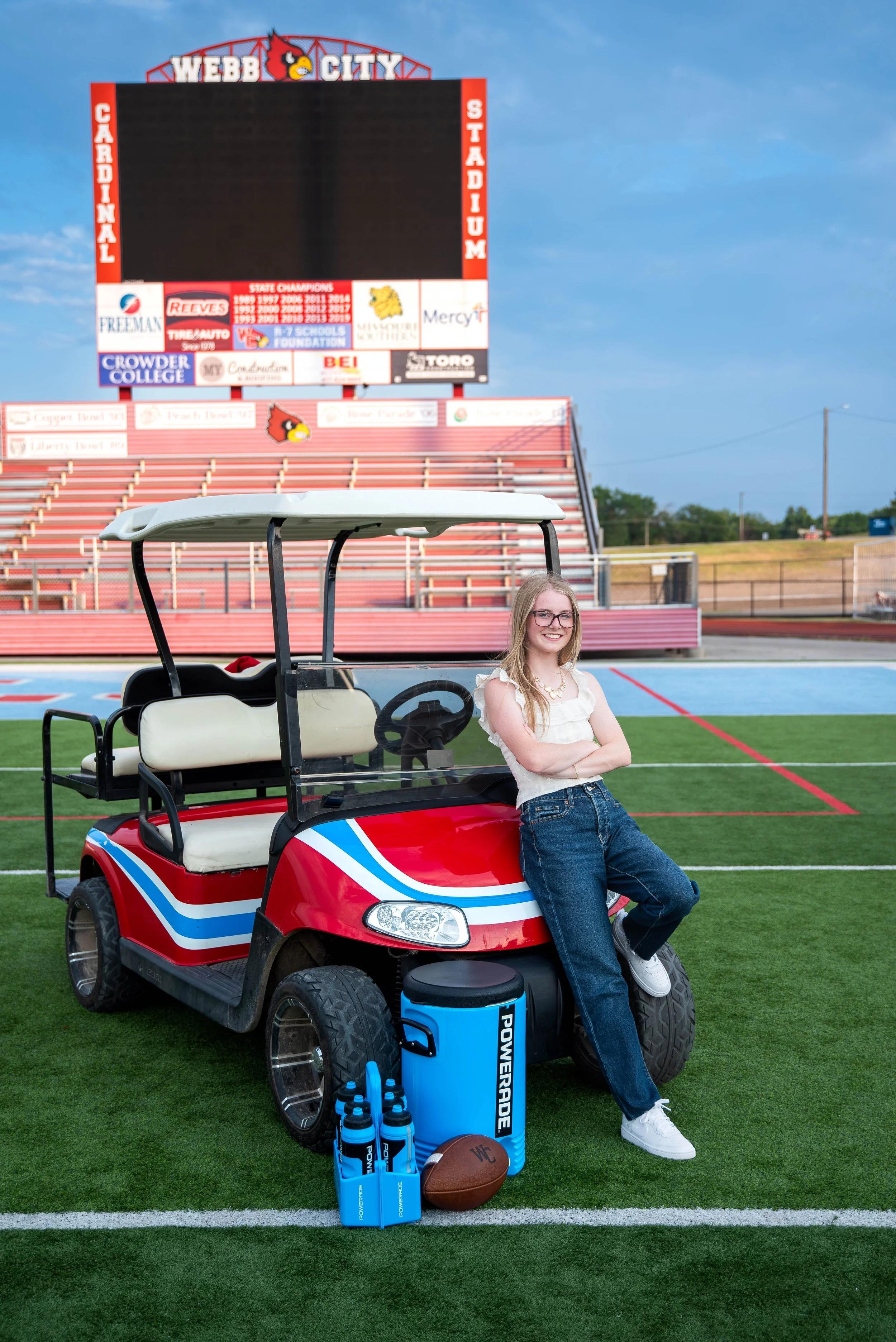 A young woman with glasses, long blonde hair, white top, and jeans, leaning against a red, white, and blue golf cart on a football field. The field has a digital scoreboard in the background that reads Webb City, surrounded by stadium seating and ban