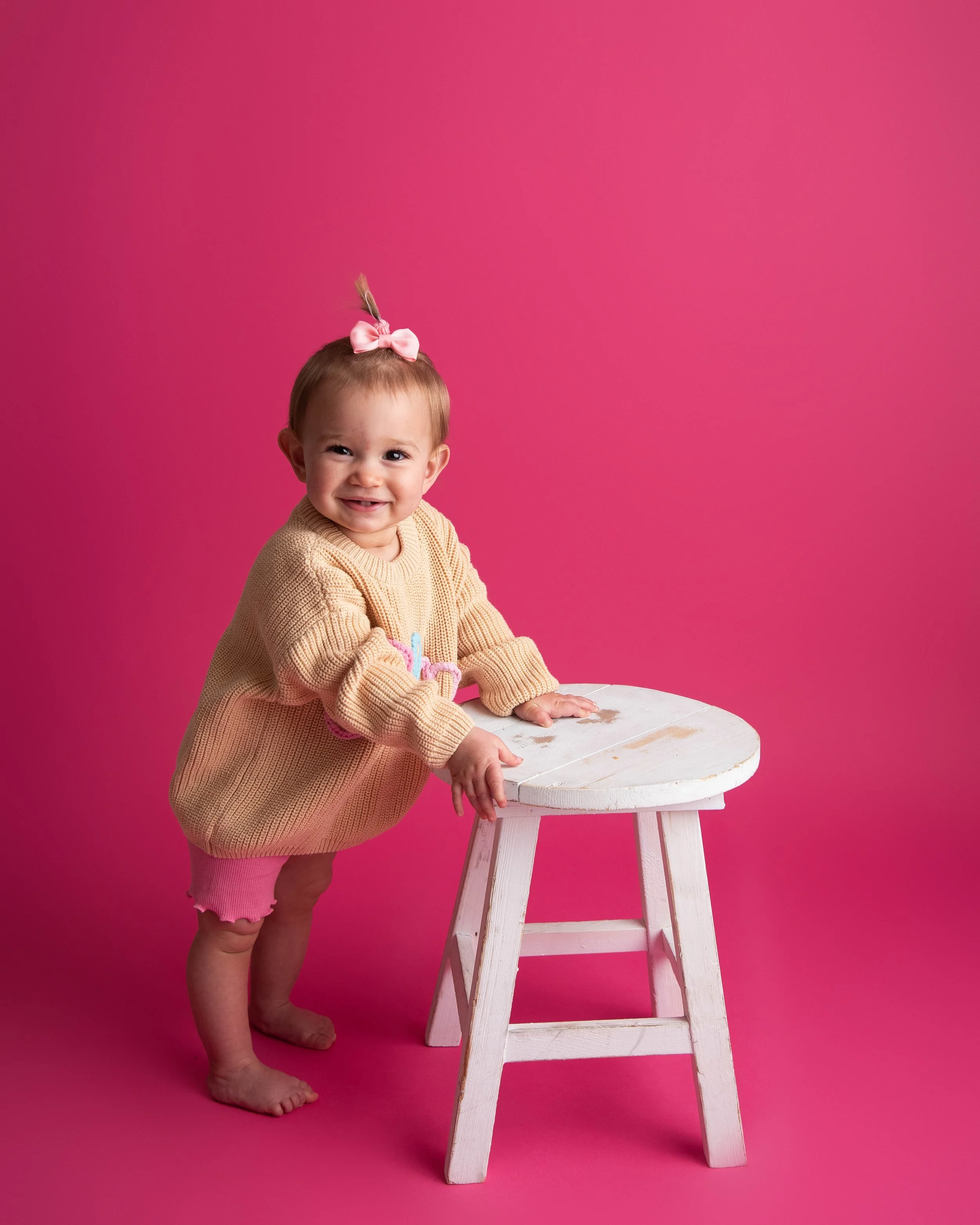 A smiling toddler girl with a pink bow in her hair, wearing a beige sweater and pink shorts, standing next to a white wooden stool against a pink background.