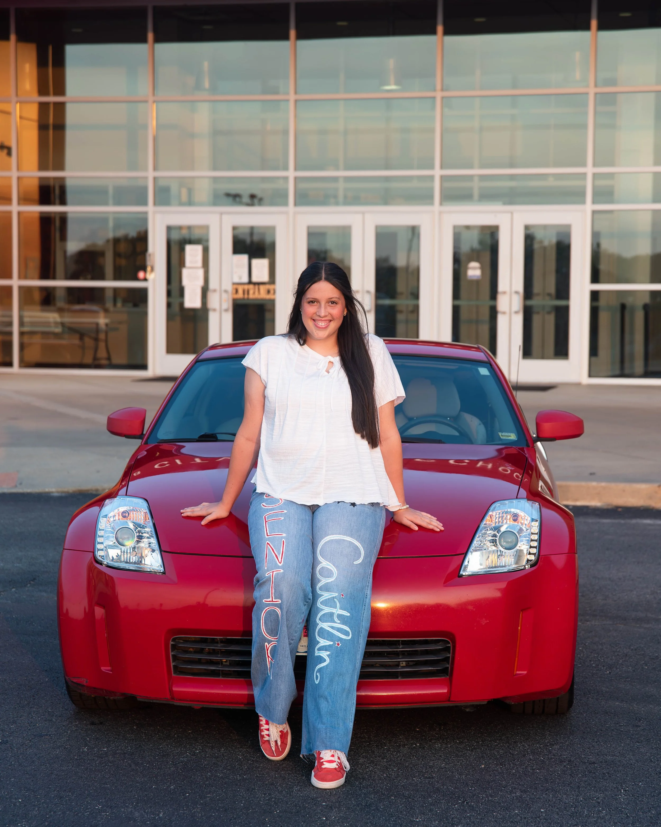 A smiling young woman with long dark hair wearing a white top and jeans with colorful writing on them, sitting on the hood of a red car in front of a modern glass building.