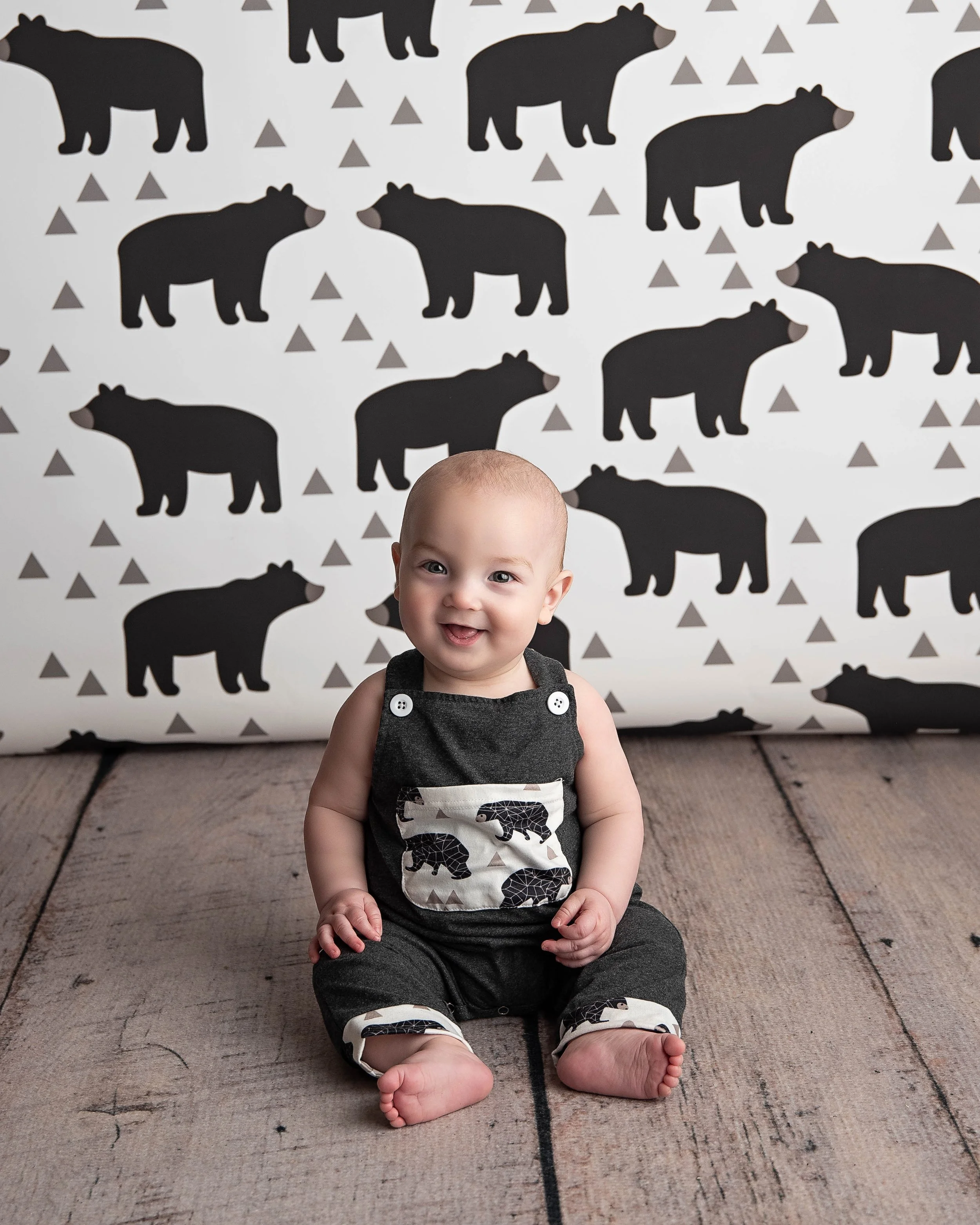 A smiling baby sitting on a wooden floor in front of a wallpaper with bear and triangle patterns, wearing a dark romper with bear-themed fabric accents.
