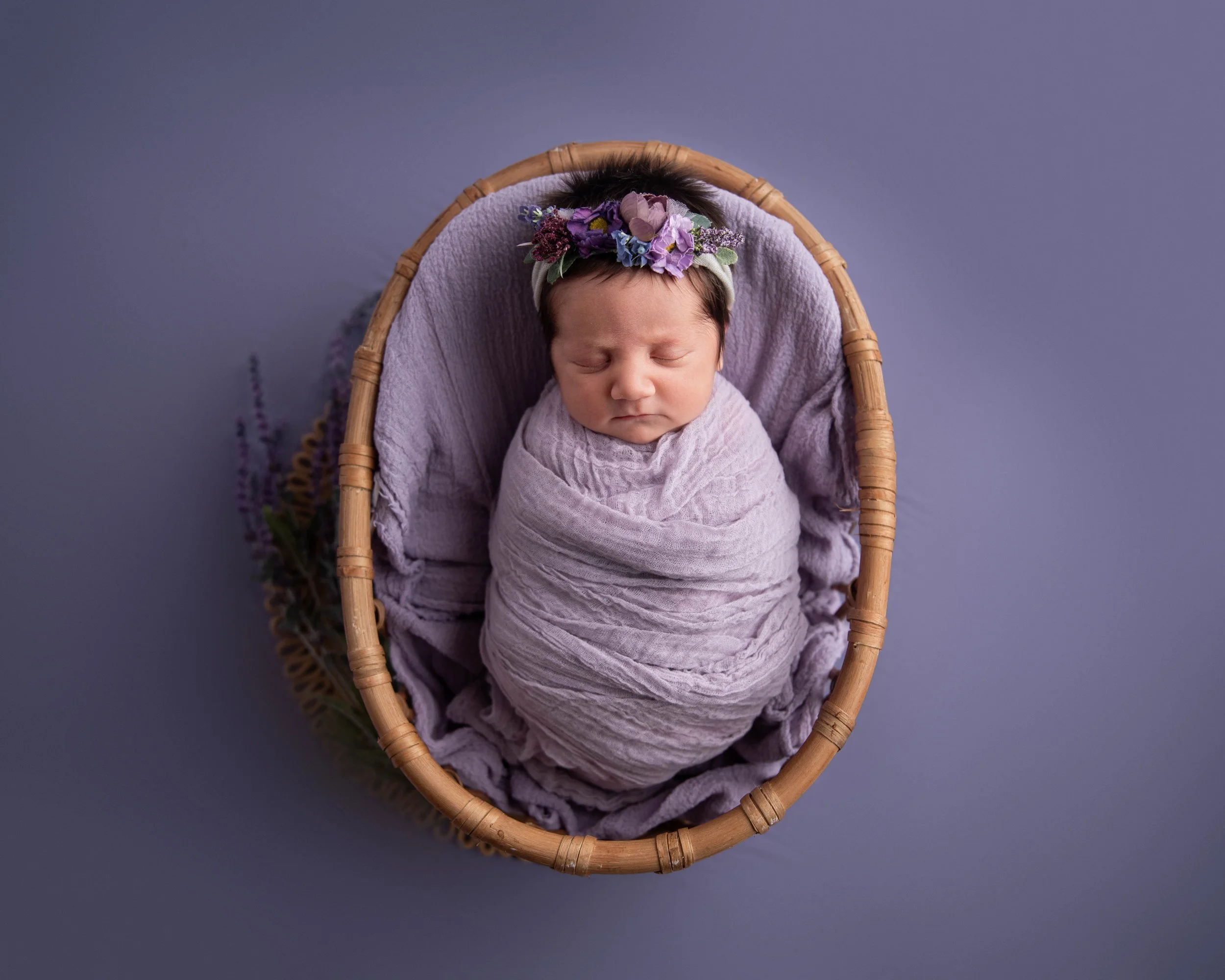 A newborn baby wrapped in a purple blanket, sleeping in a round wicker basket decorated with purple flowers and greenery.