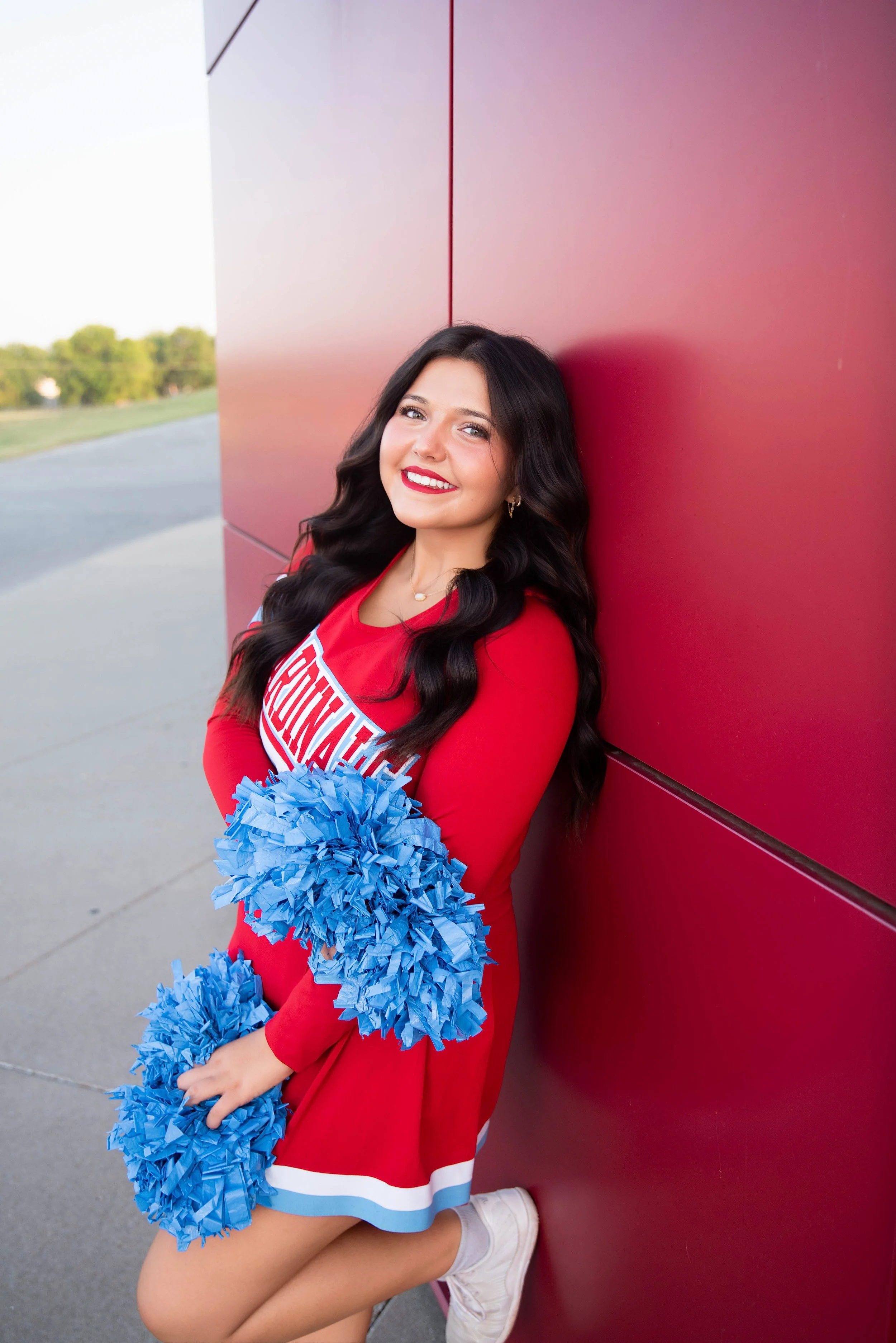 A young woman dressed as a cheerleader with a red uniform and blue pom-poms, smiling and leaning against a red wall outdoors.