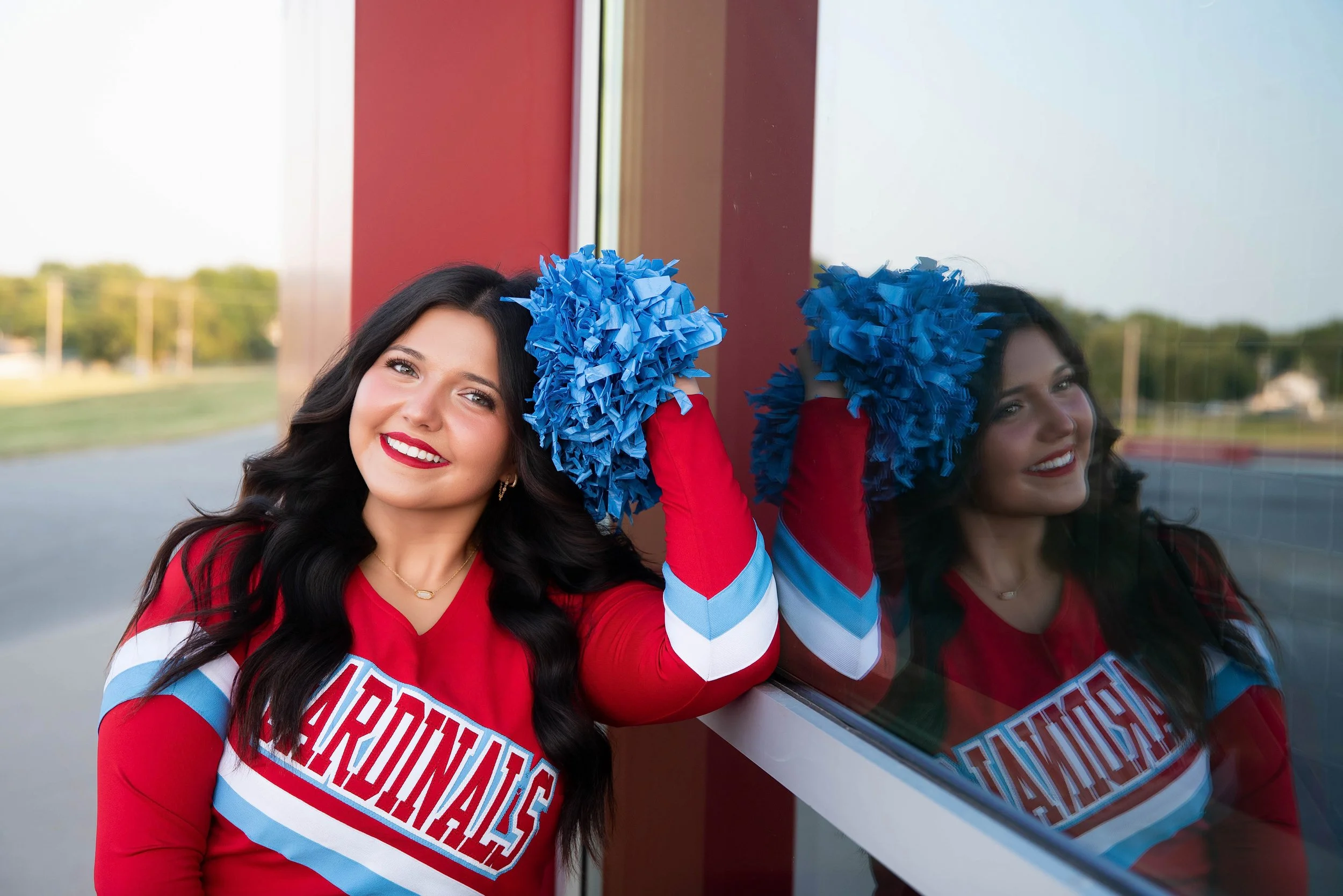 A young woman wearing a red cheerleading uniform with 'Cardinals' on the front, holding blue pom-poms, smiling, standing next to a reflective glass wall that shows her reflection.