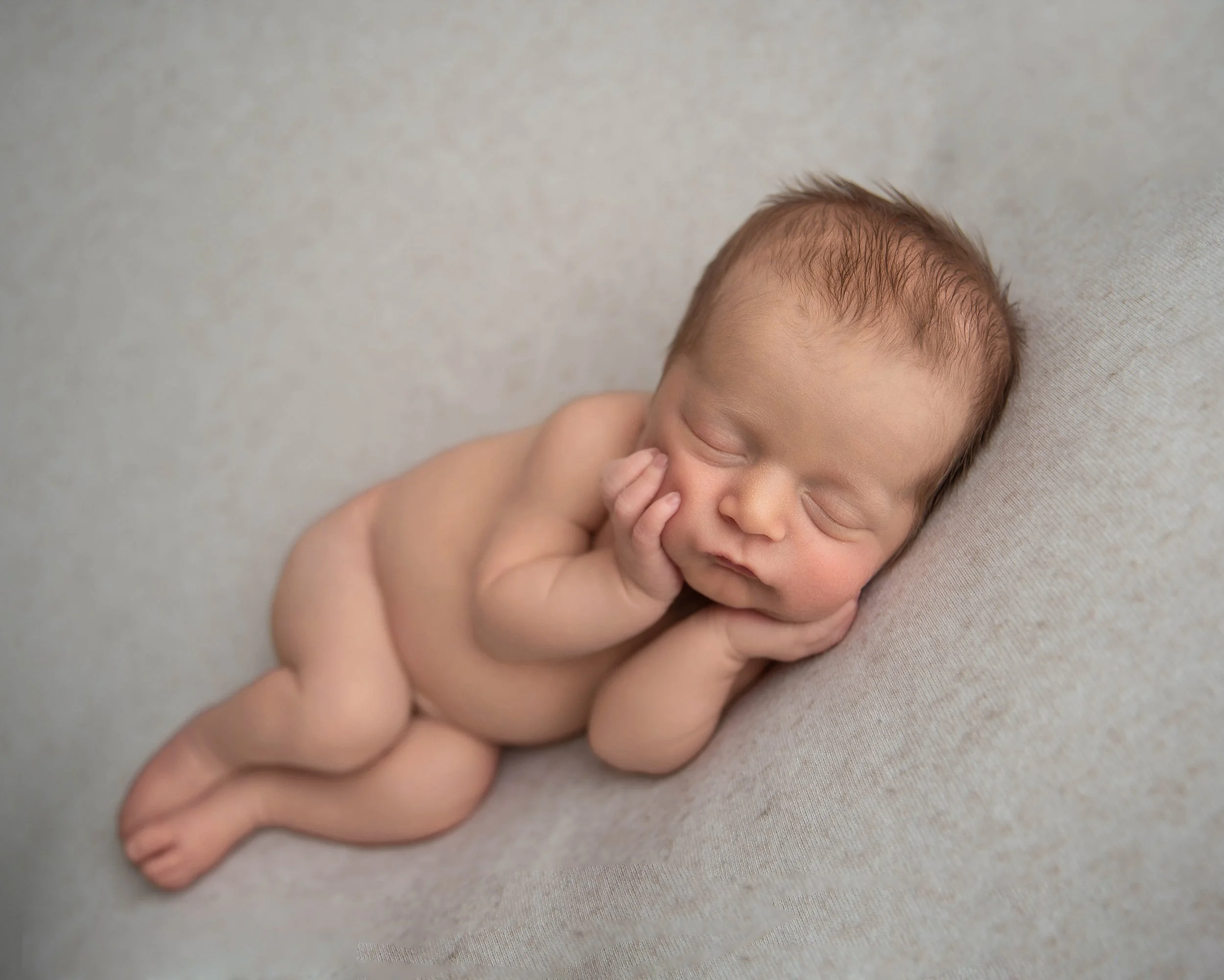 A sleeping newborn baby lying on a soft surface, resting on its side with hands near face.