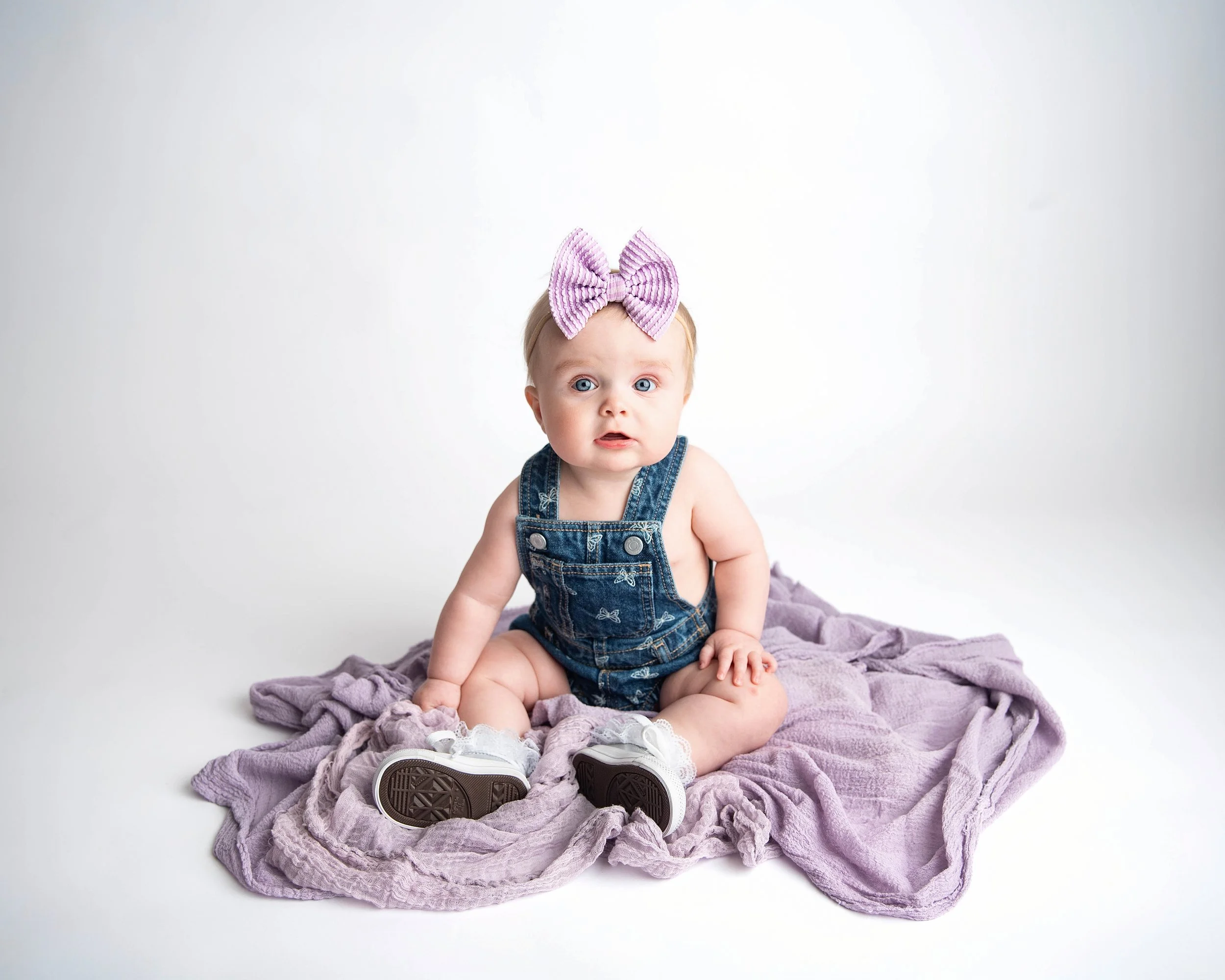 A baby girl with blue eyes sitting on a purple blanket against a white background, wearing denim overalls, a pink bow headband, and white shoes.