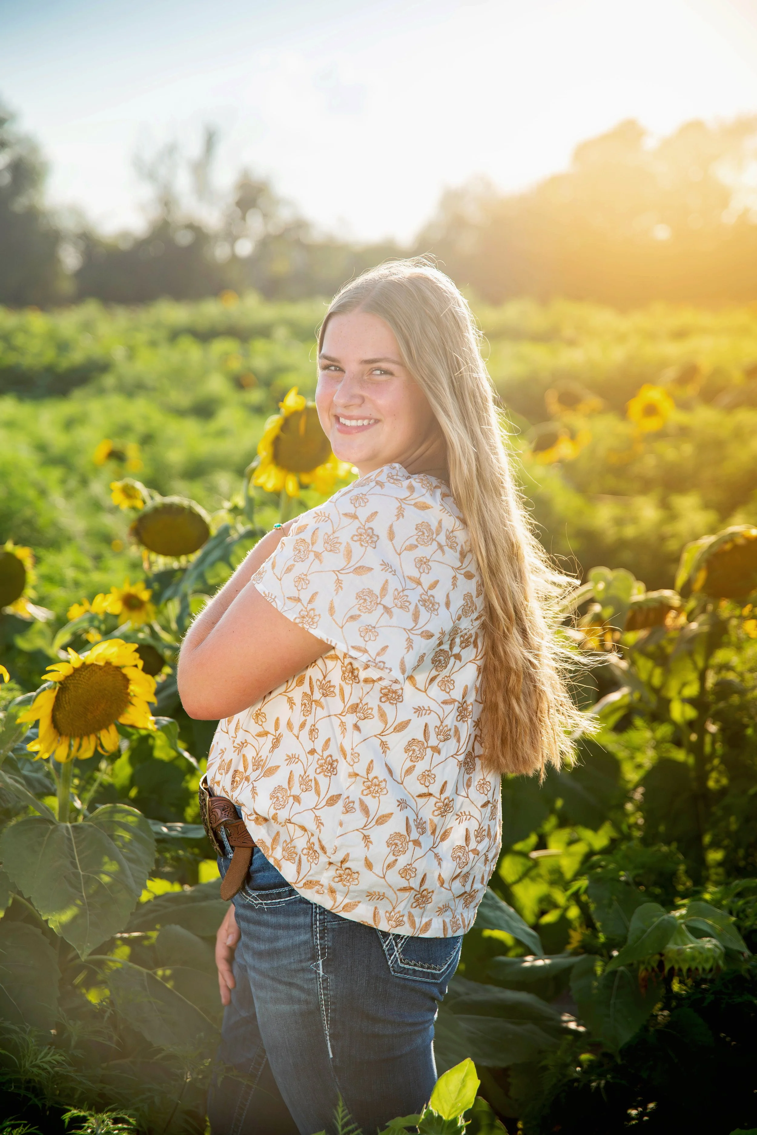 A young woman with long blonde hair smiling in a sunflower field at sunset.