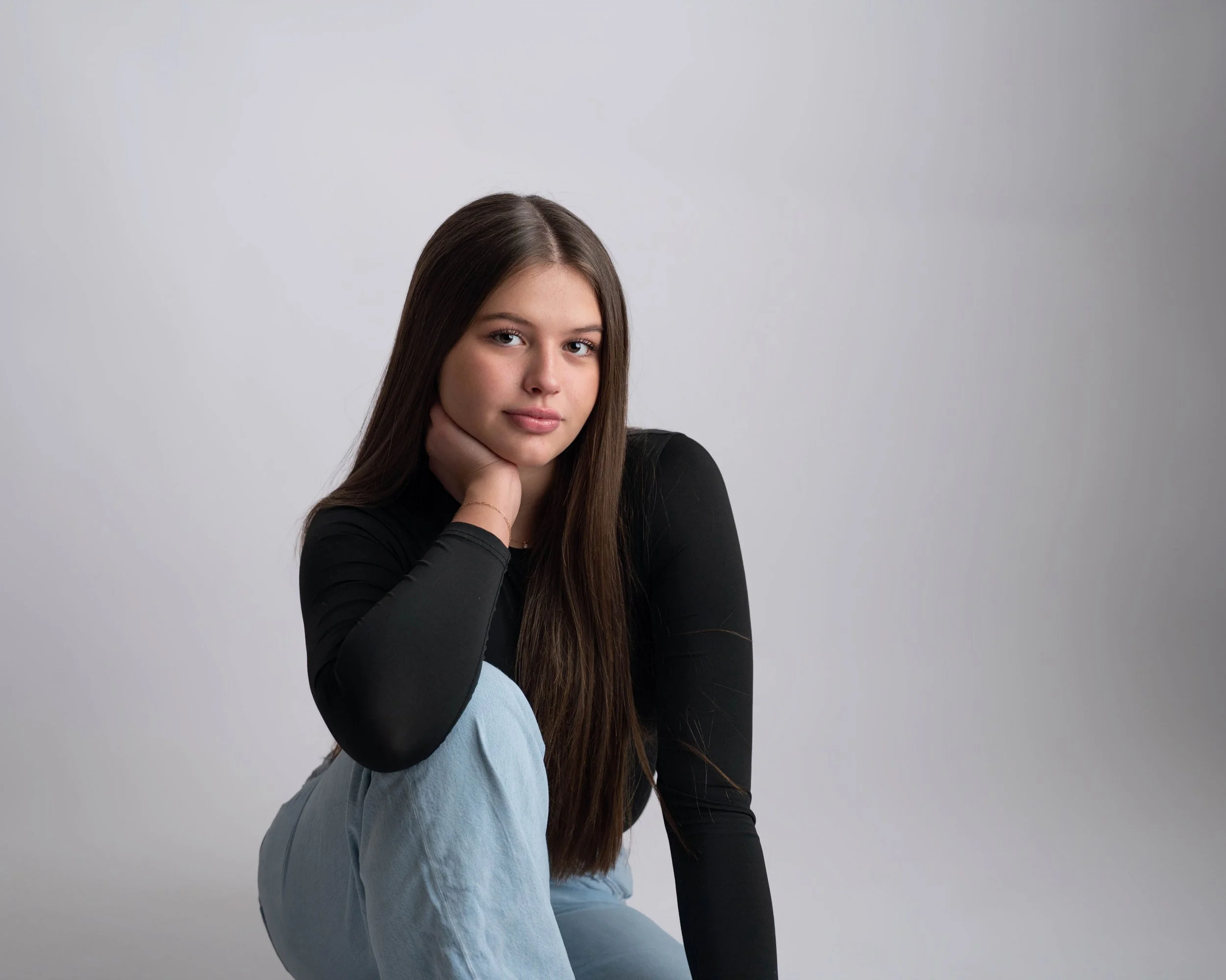 A young woman with long brown hair wearing a black long sleeve top and light blue jeans, sitting against a plain gray background, with her chin resting on her hand and looking at the camera.