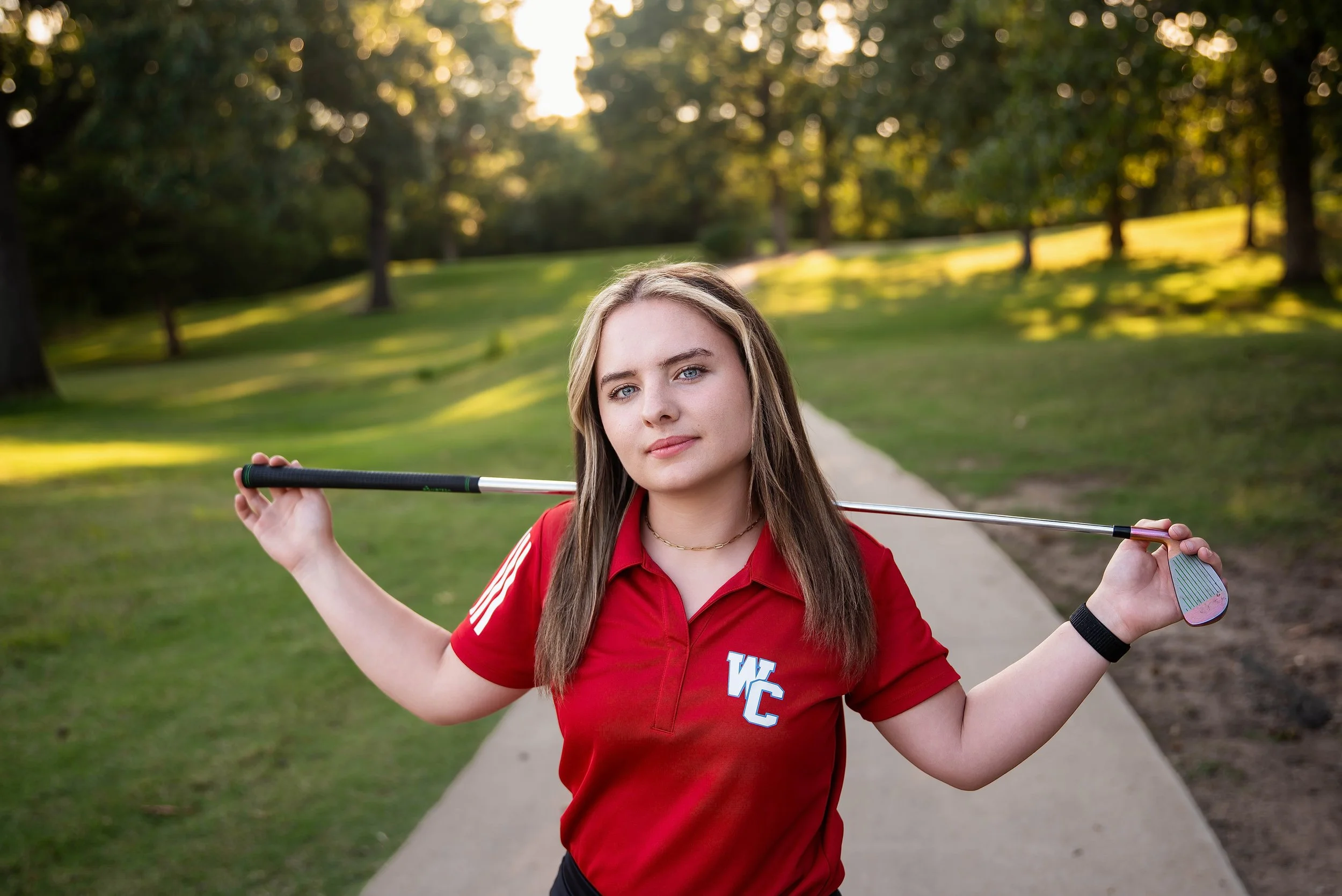 A young woman in a red sports shirt holding a golf club across her shoulders on a golf course during sunset.
