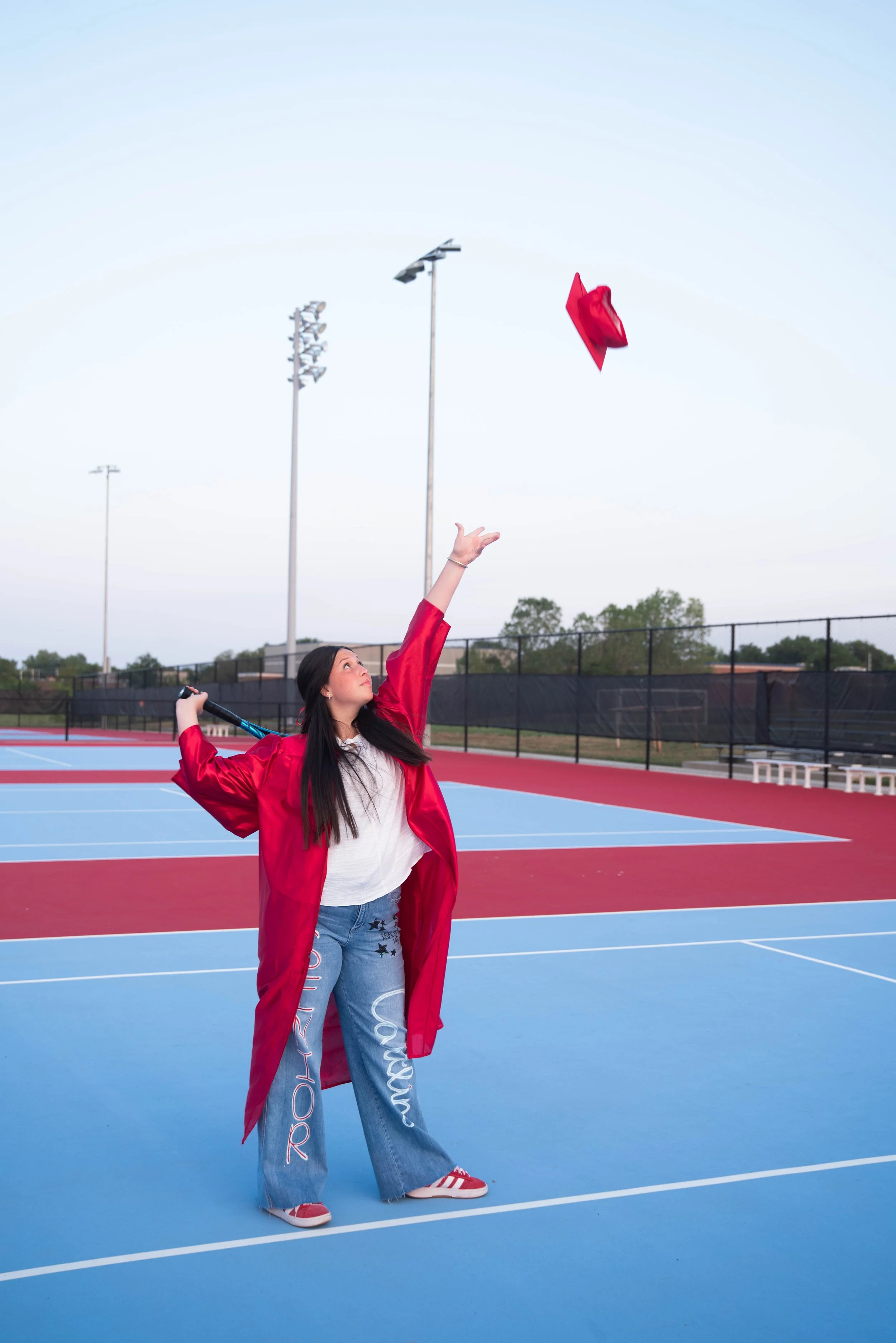 A young woman in a red graduation gown and jeans is on a tennis court, tossing her cap into the air.