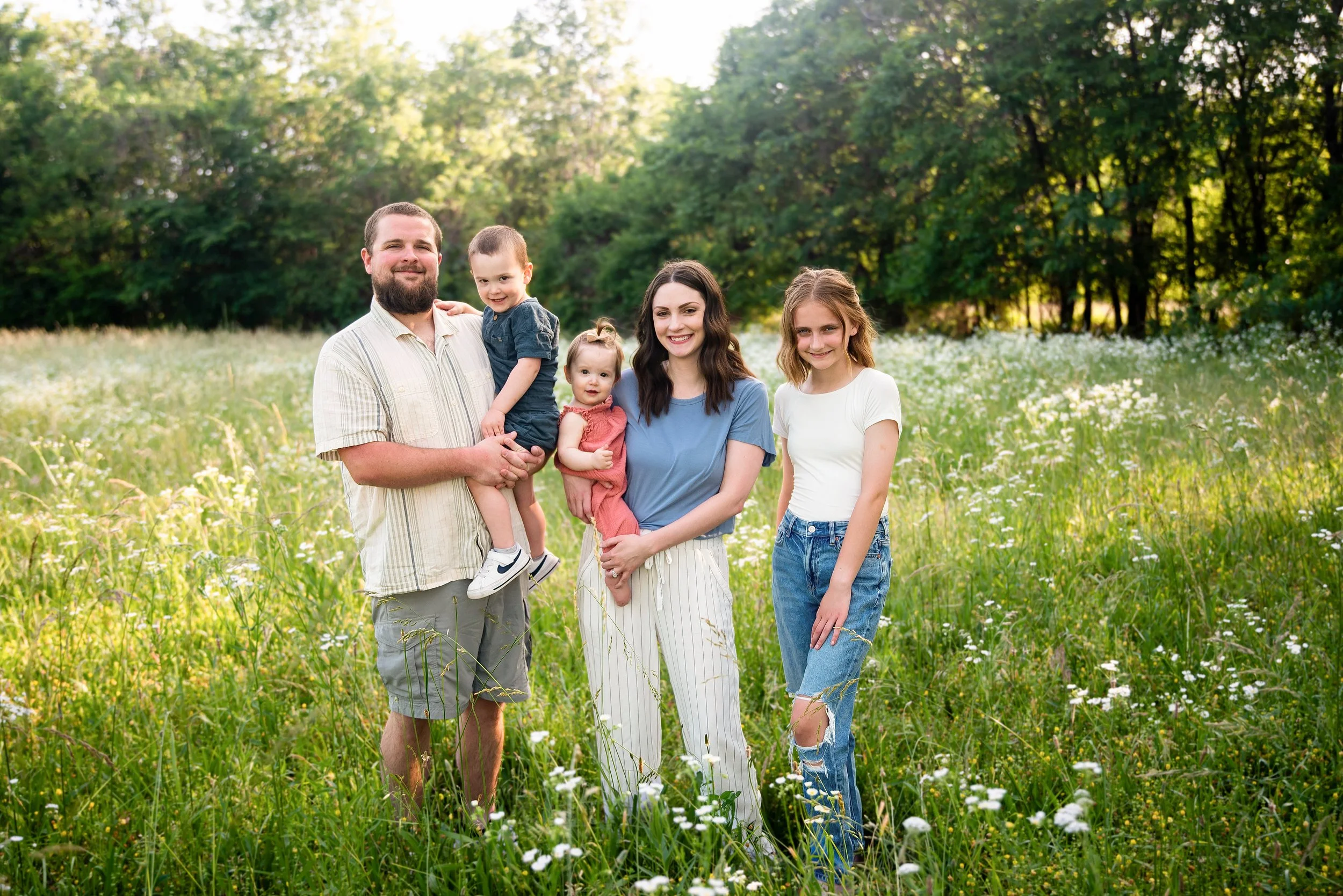 A family of five standing in a grassy field with wildflowers and trees in the background during daylight. The man is holding a young boy, and two young girls are standing beside a woman with dark hair.