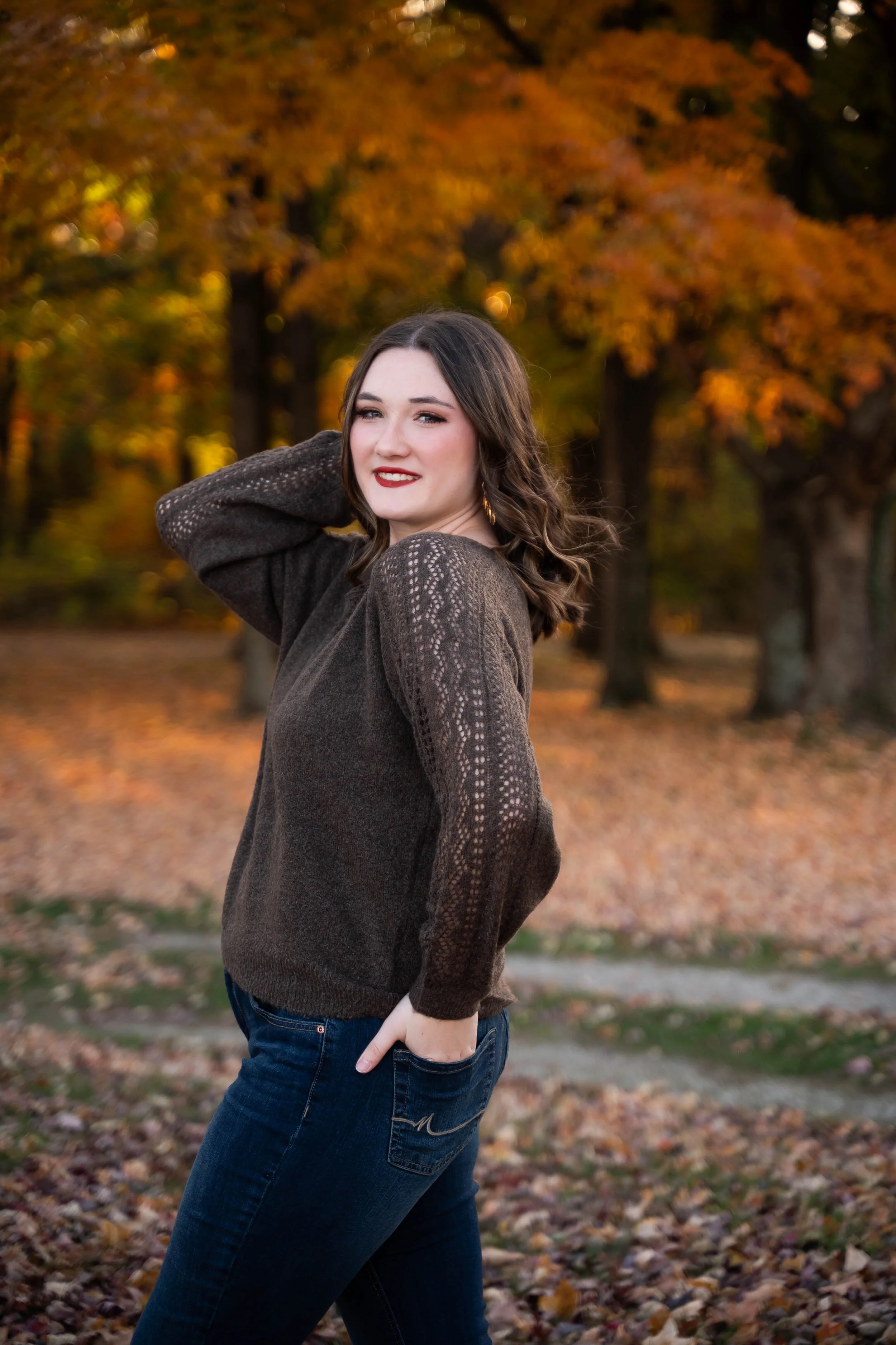 A woman with wavy brown hair, wearing a brown knit sweater and dark jeans, standing outdoors in fall setting with colorful trees and fallen leaves, smiling at the camera.
