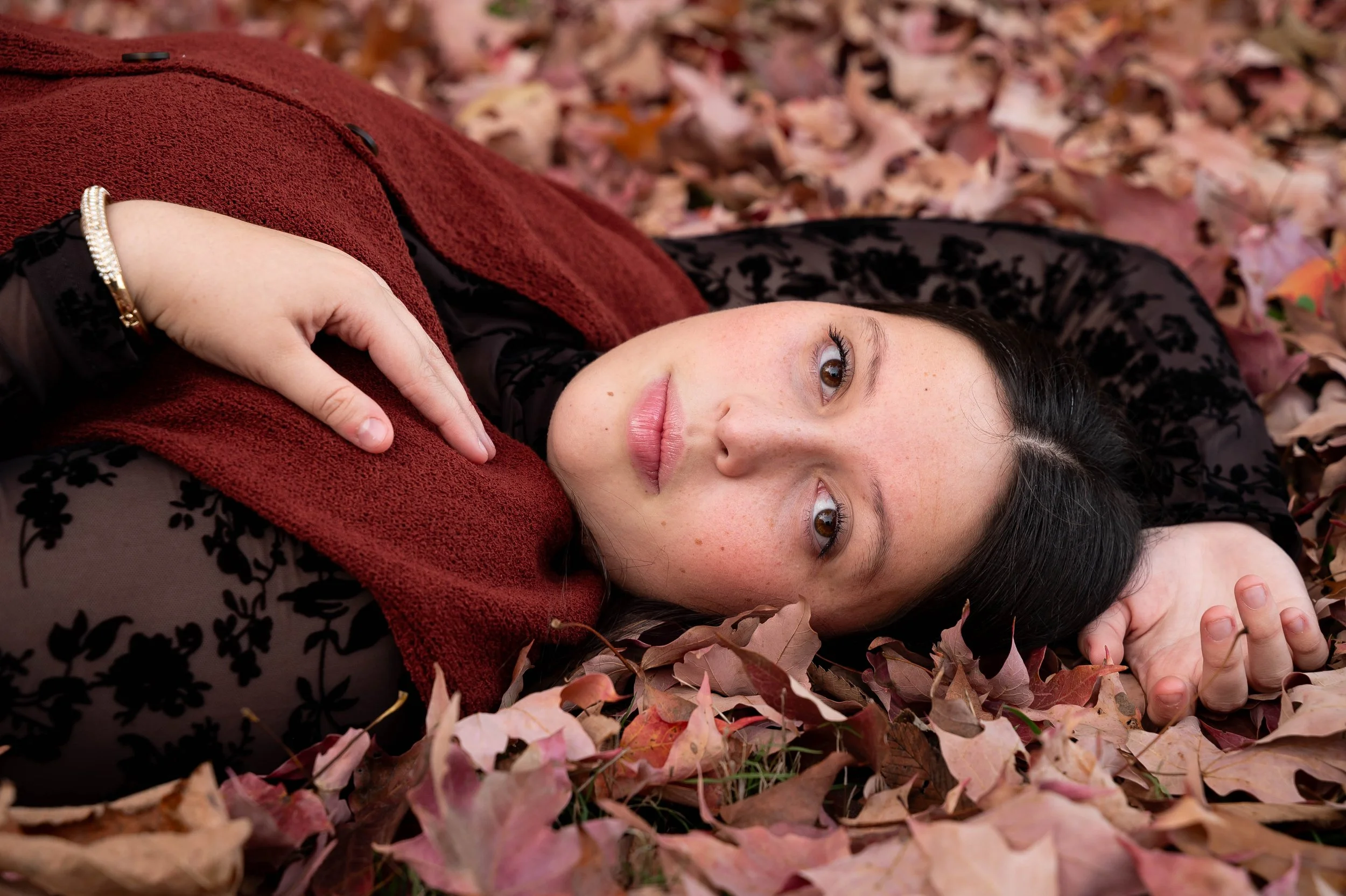 A young woman with dark hair and freckles lies on a bed of fallen autumn leaves, gazing softly at the camera. She wears a black lace top underneath a reddish-brown textured vest and has a bracelet on her wrist.