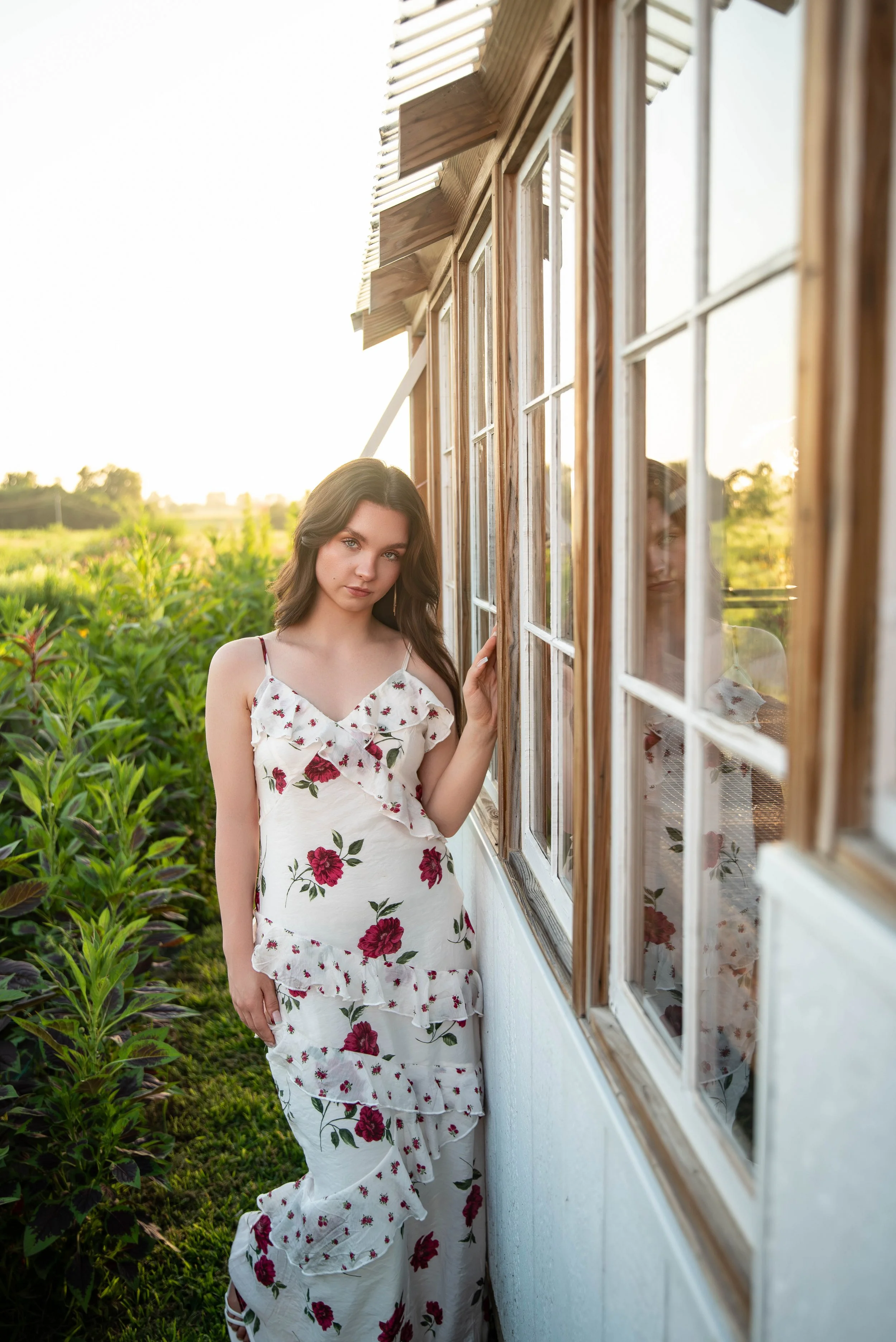 A young woman in a white dress with red and pink floral patterns standing beside a wooden barn with glass windows, outdoors during sunset with green plants in the background.