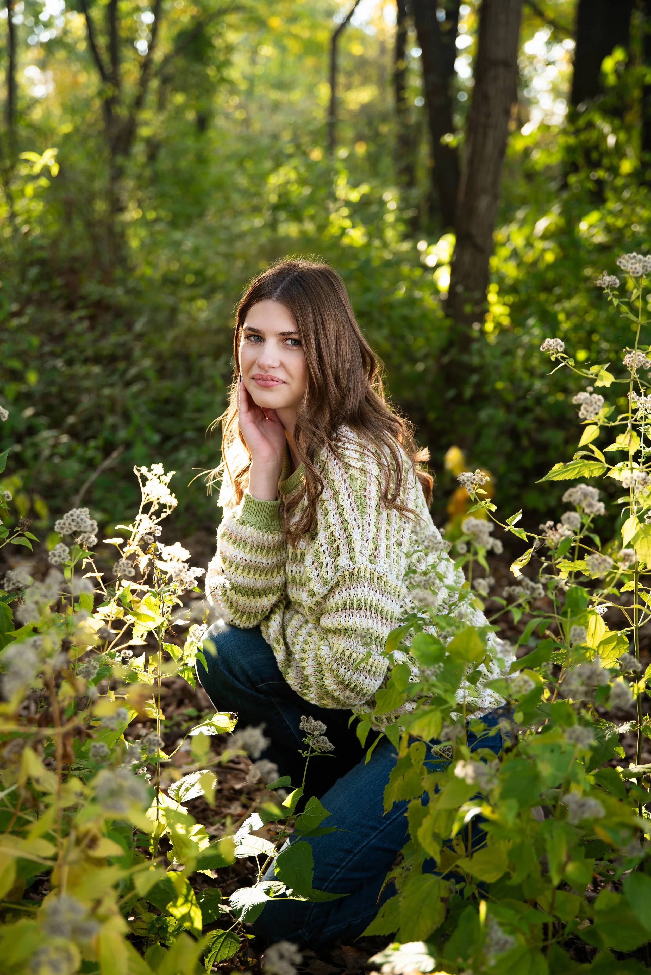 Young woman with long brown hair kneeling in a forested area surrounded by green plants and white flowers, wearing a striped sweater and jeans, looking at the camera.