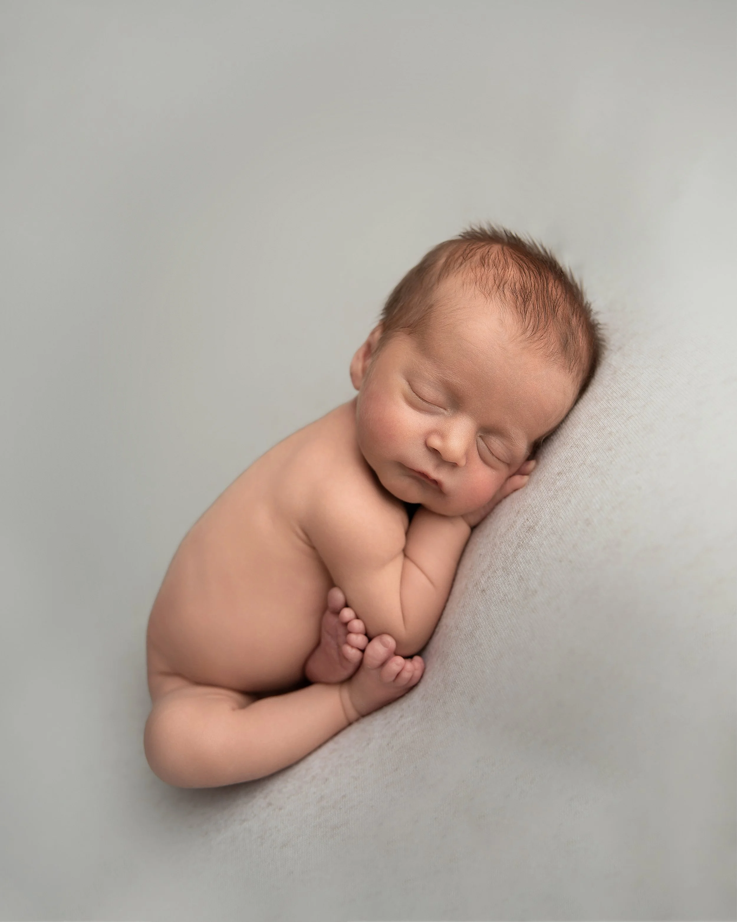 A sleeping newborn baby with a soft skin tone, lying on its side on a light-colored surface, with its hands gently tucked under its cheek and legs curled up.