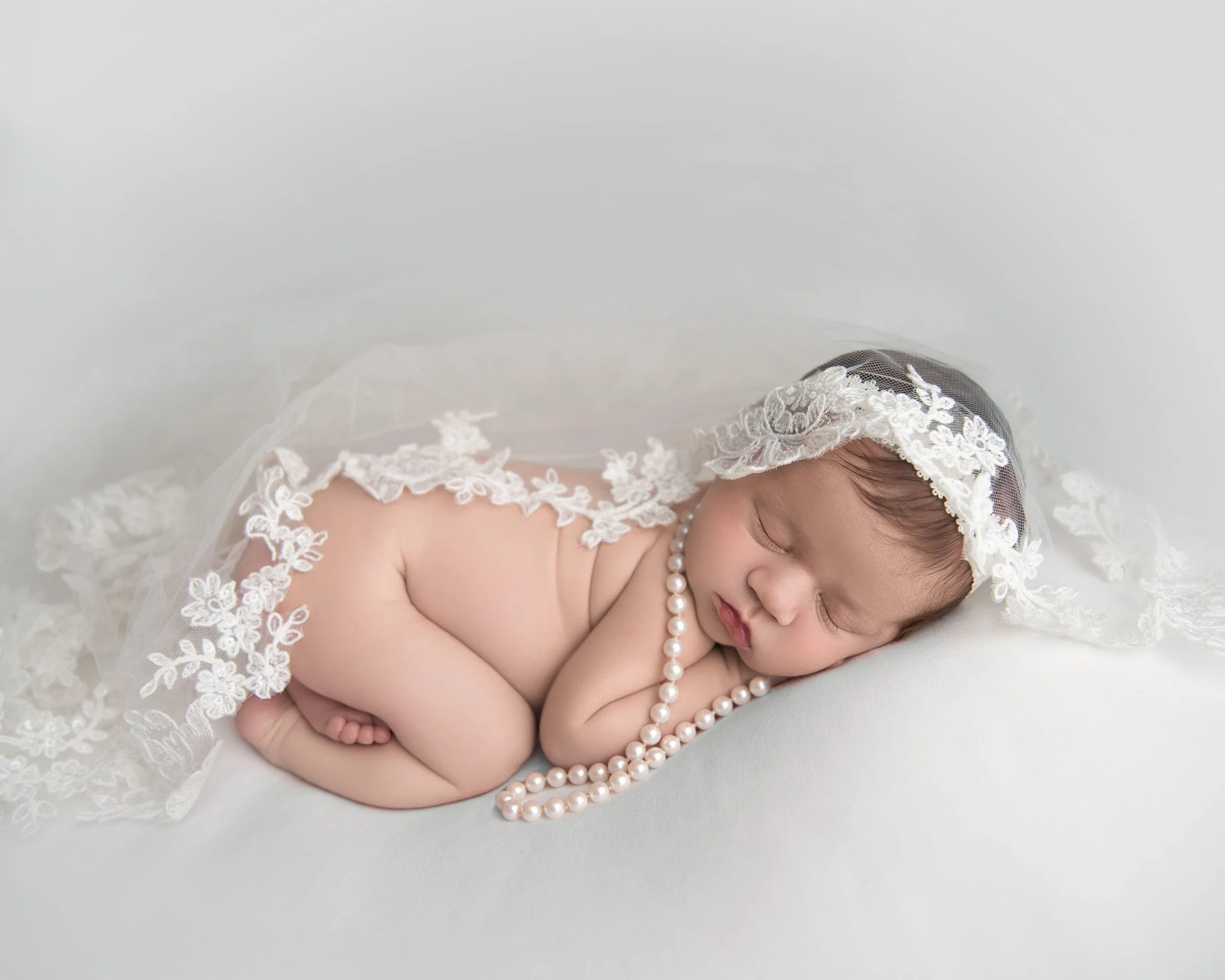 A sleeping baby girl wearing a lace veil, pearl necklace, and lying on a soft white surface.
