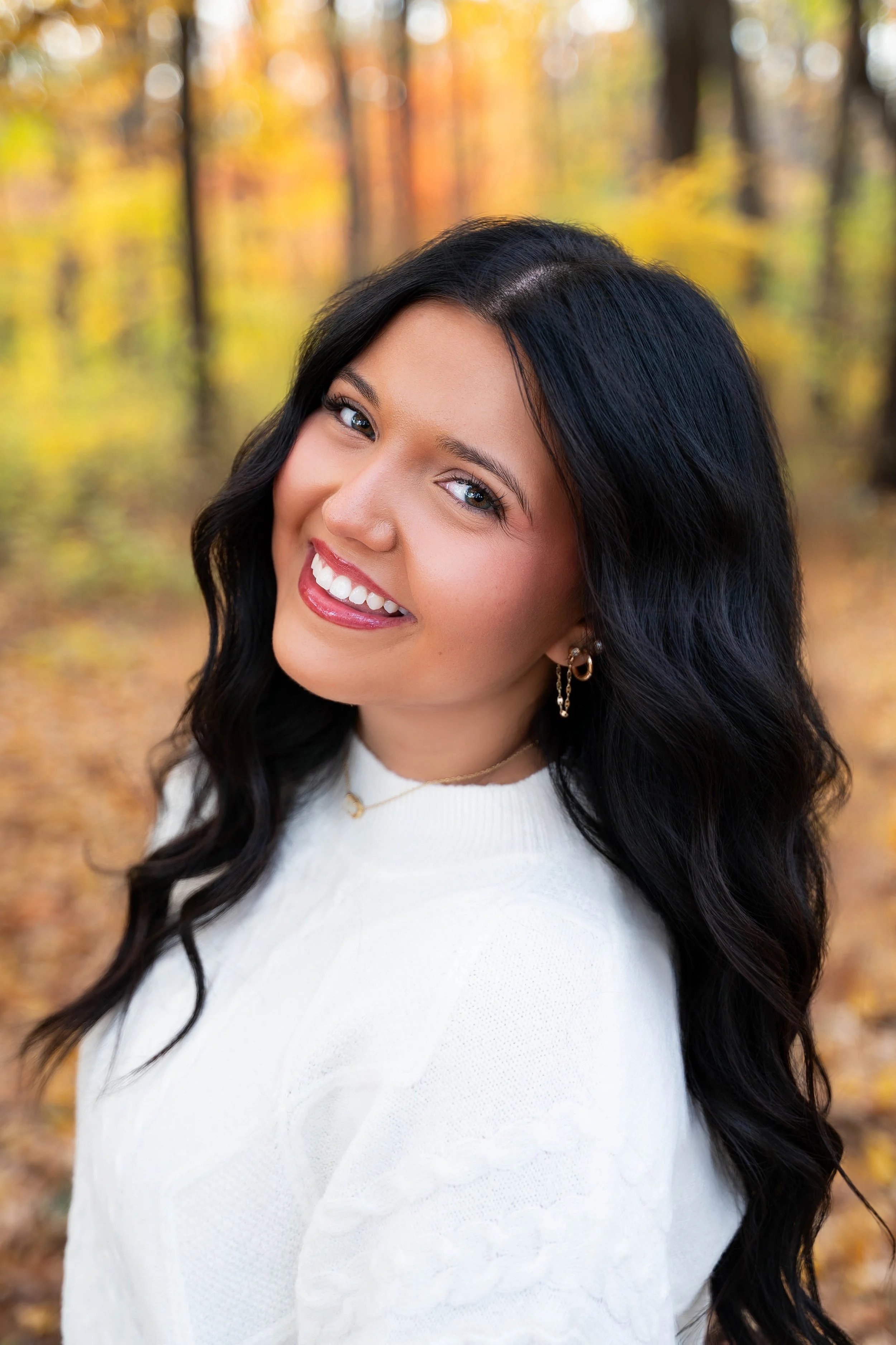 A smiling woman with long black hair wearing a white sweater standing outdoors in an autumn forest with yellow and orange leaves.