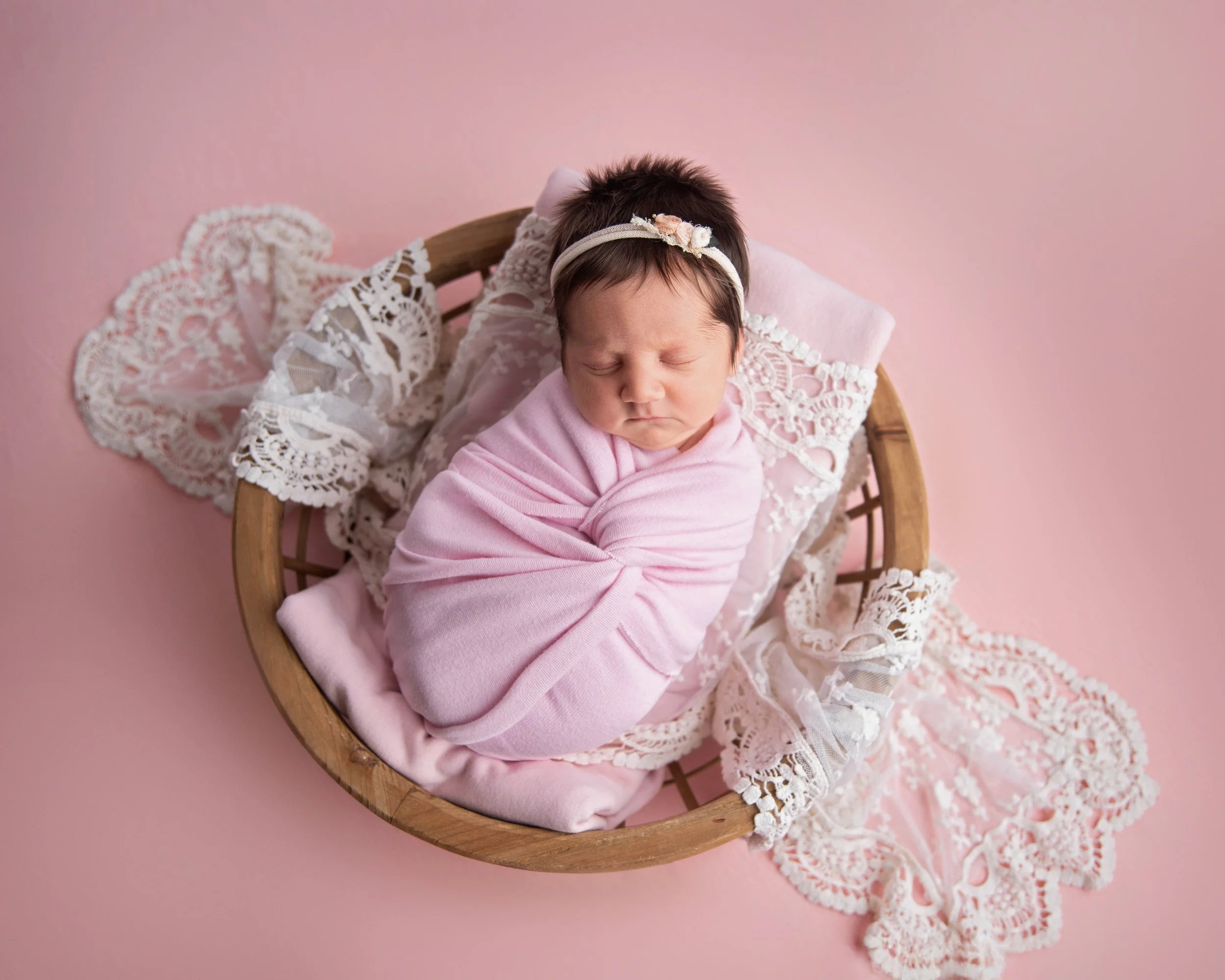 A newborn baby girl sleeping swaddled in a pink blanket, lying on a pink surface with a white lace doily and a pink cloth inside a round wooden basket.