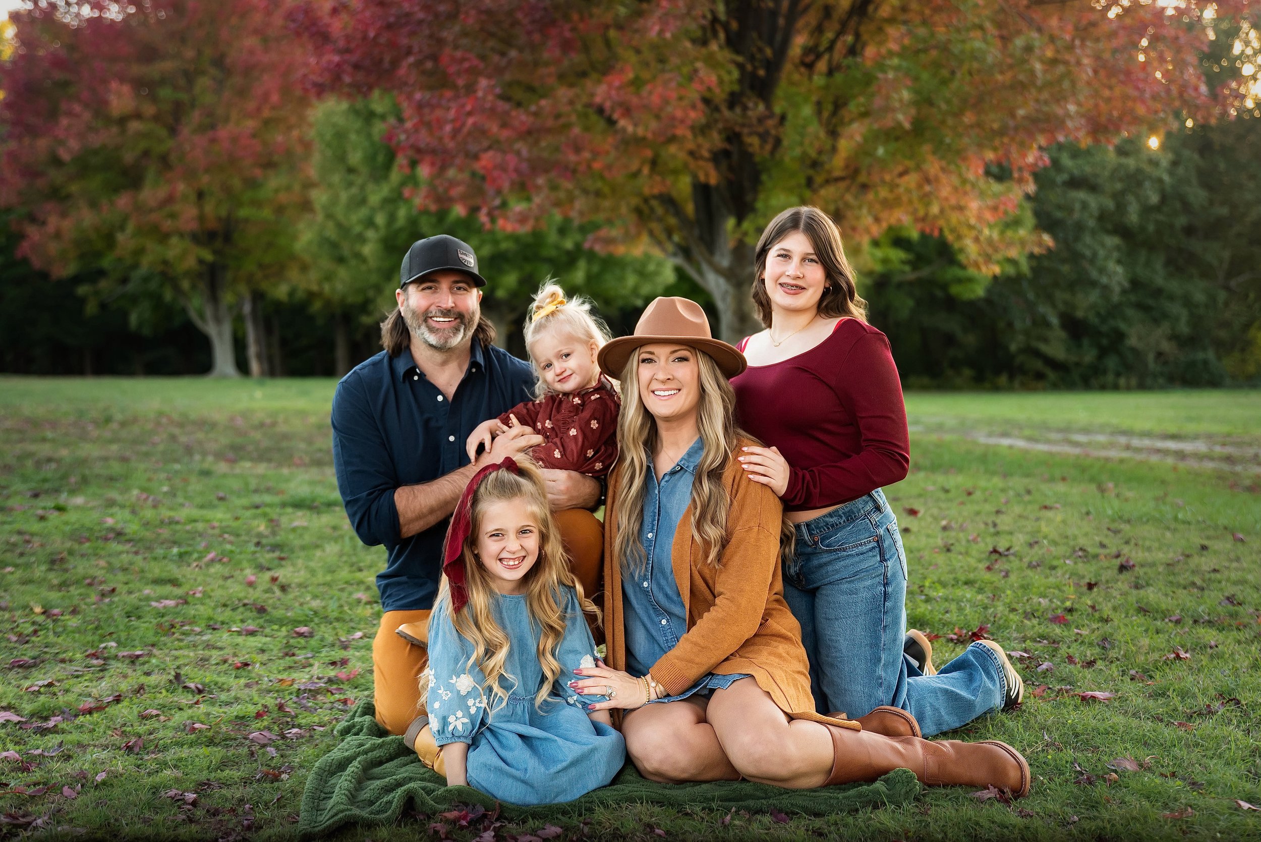 A family of five posing on a grassy field with colorful autumn trees in the background.