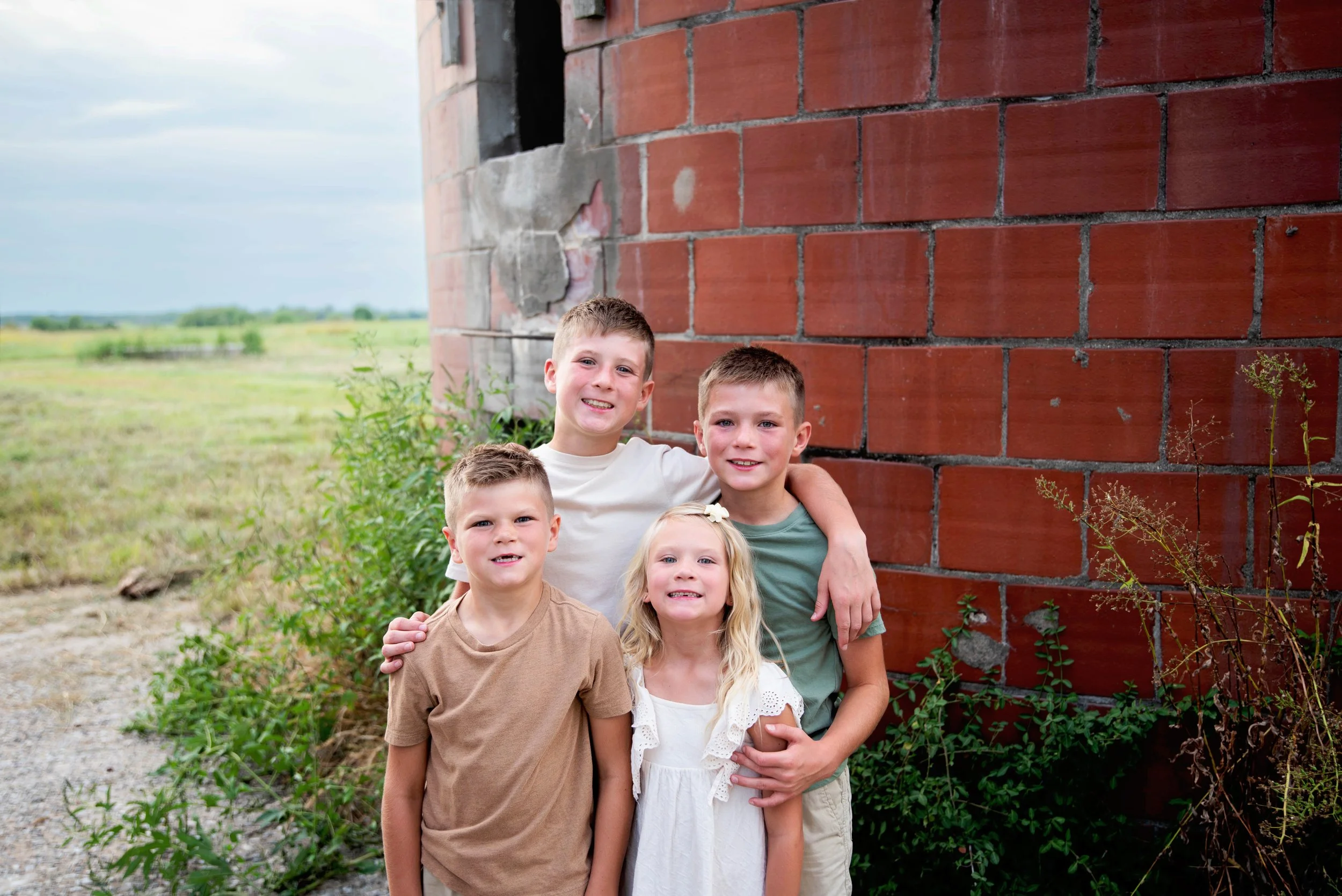 Group of four children standing outdoors near a brick building with greenery in the background.