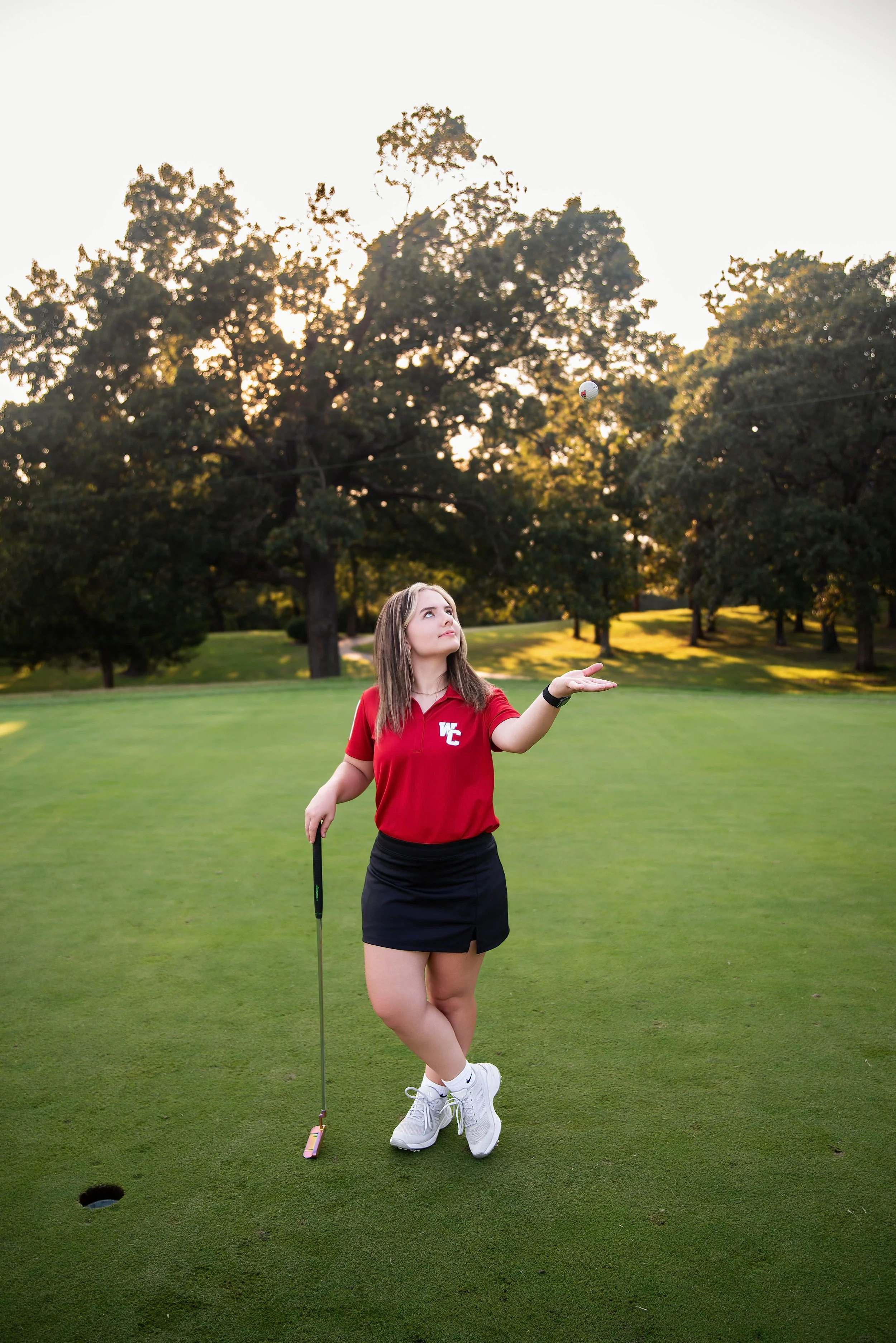 A young woman in a red golf shirt and black skirt on a golf course, appears to be playing golf, with a golf ball in the air and a hole with a flag nearby.