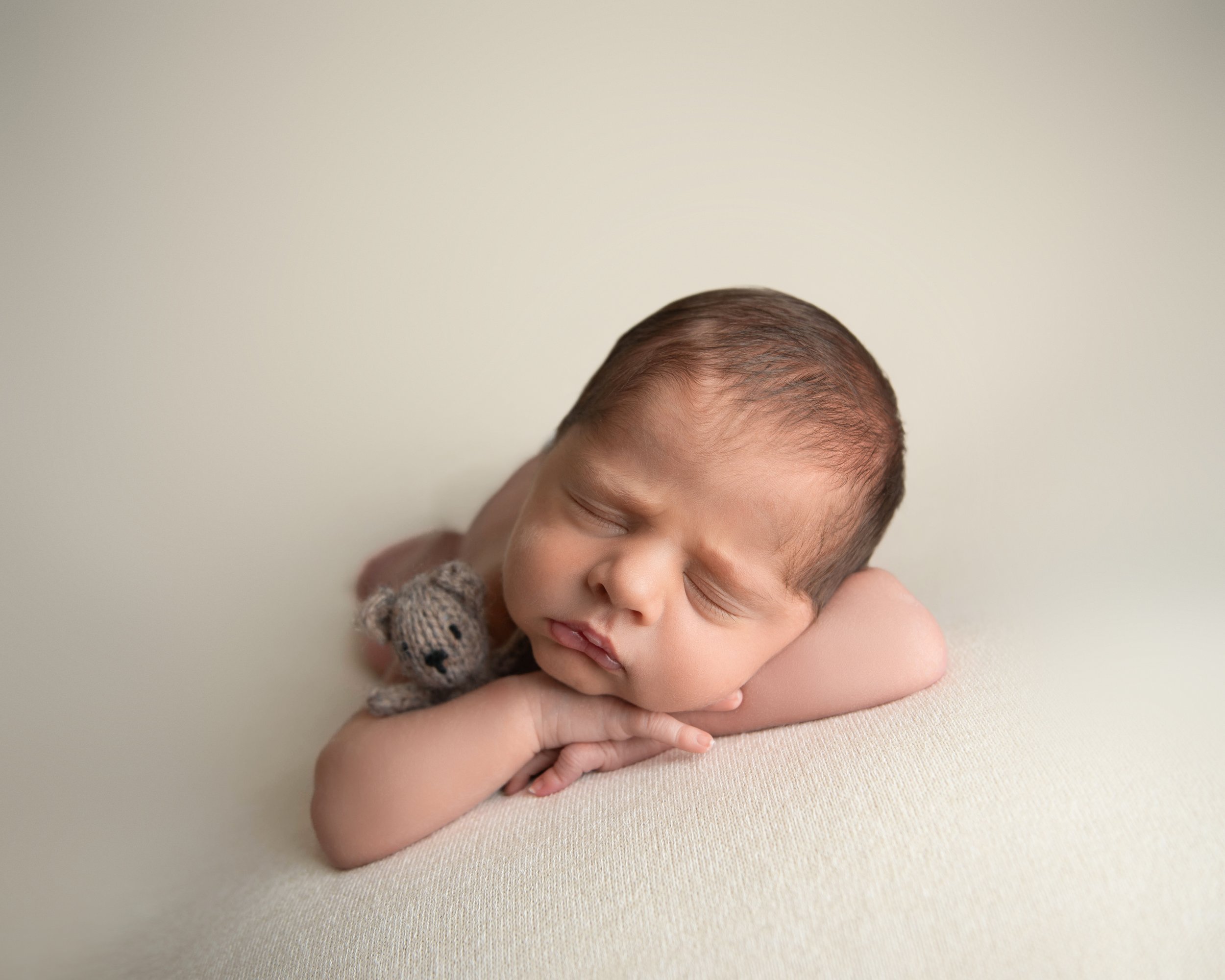 A newborn baby sleeping peacefully with a small knit teddy bear on his shoulder, resting on a soft beige surface.
