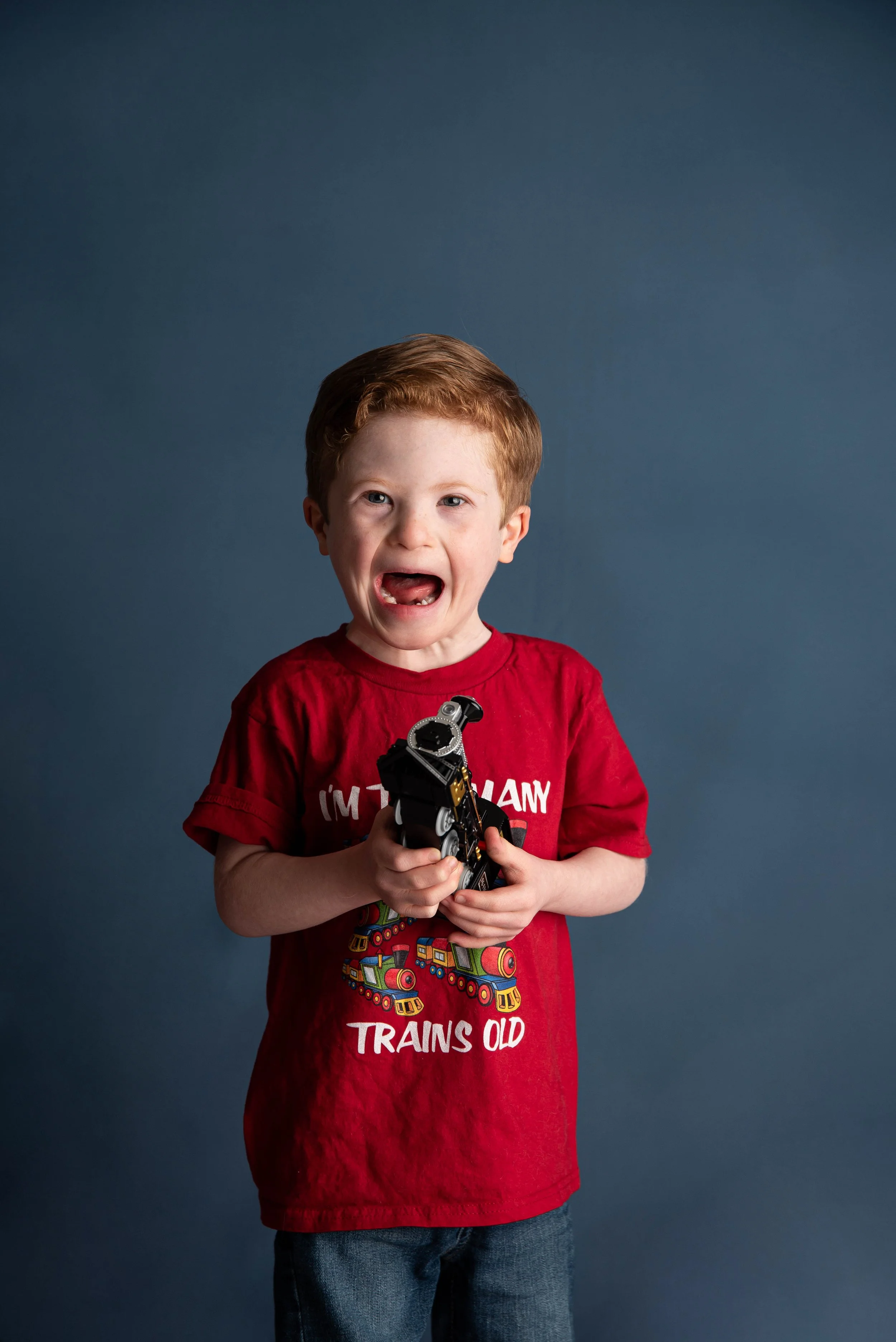 A young boy with red hair wearing a red T-shirt with a train graphic and text, holding a small toy train, standing against a plain dark background.
