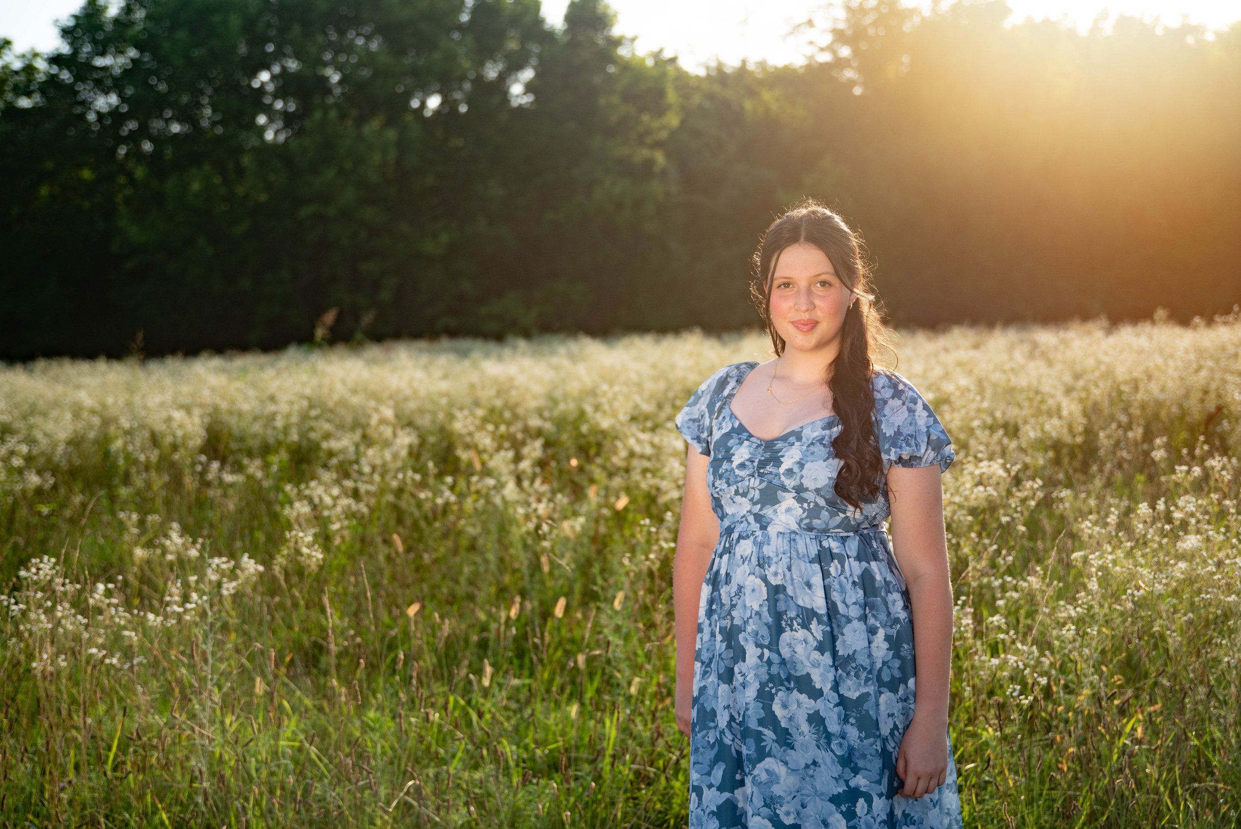 A young woman in a blue floral dress standing in a field of white flowers during sunset.