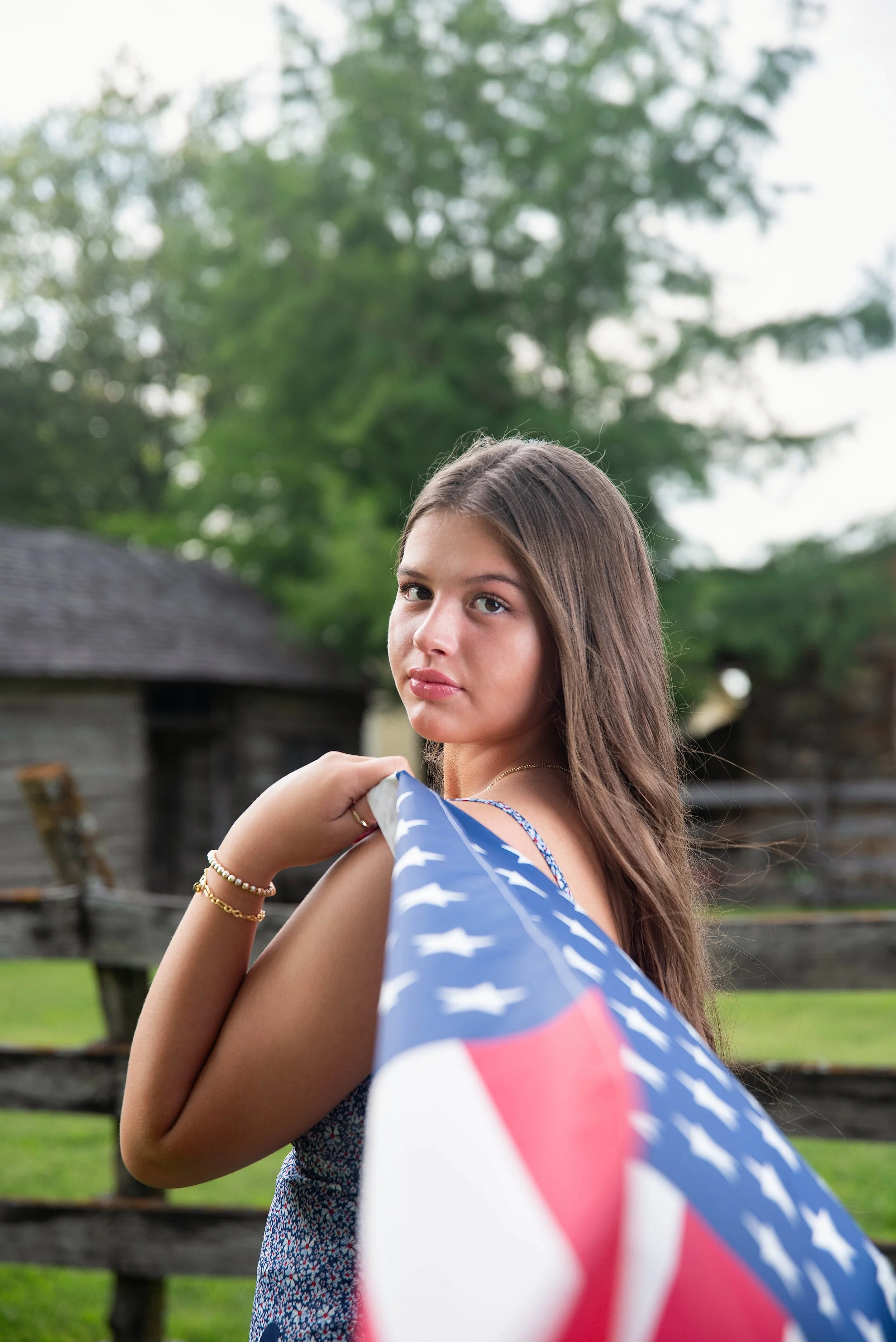 Young woman with long brown hair holding an American flag outdoors, with a rustic wooden fence and green trees in the background.