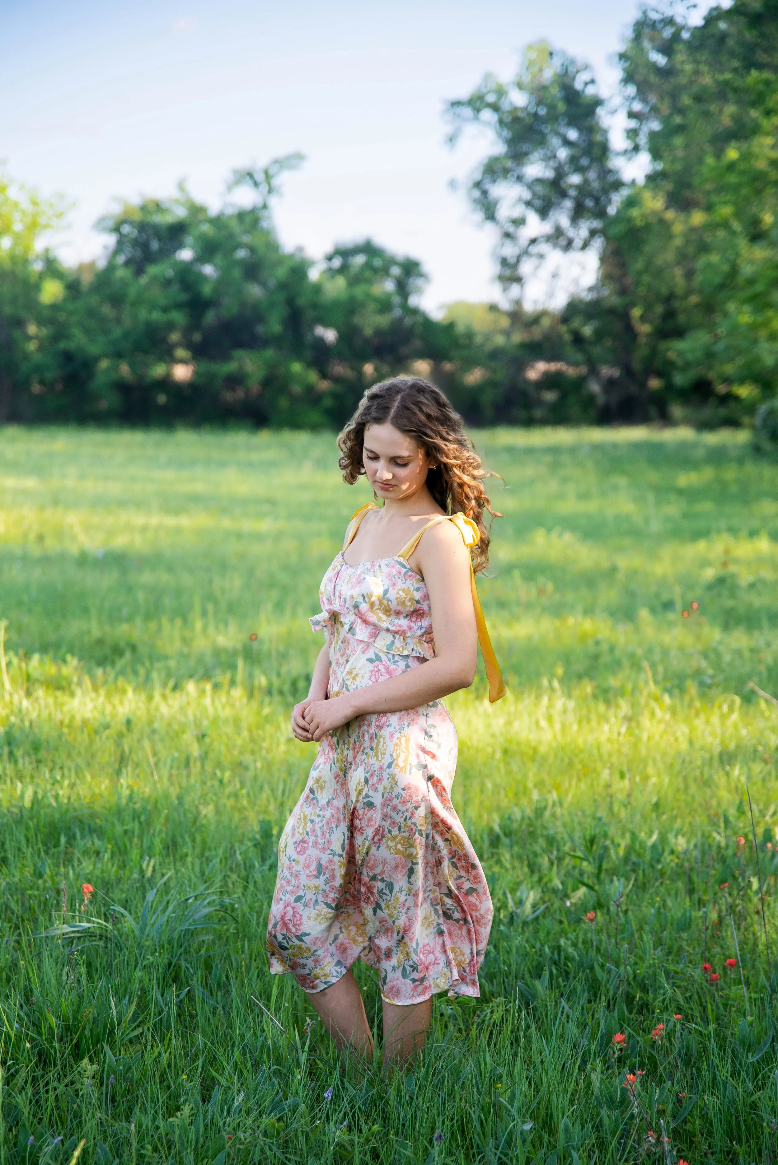 Young woman in floral dress standing in a green meadow with trees in the background.