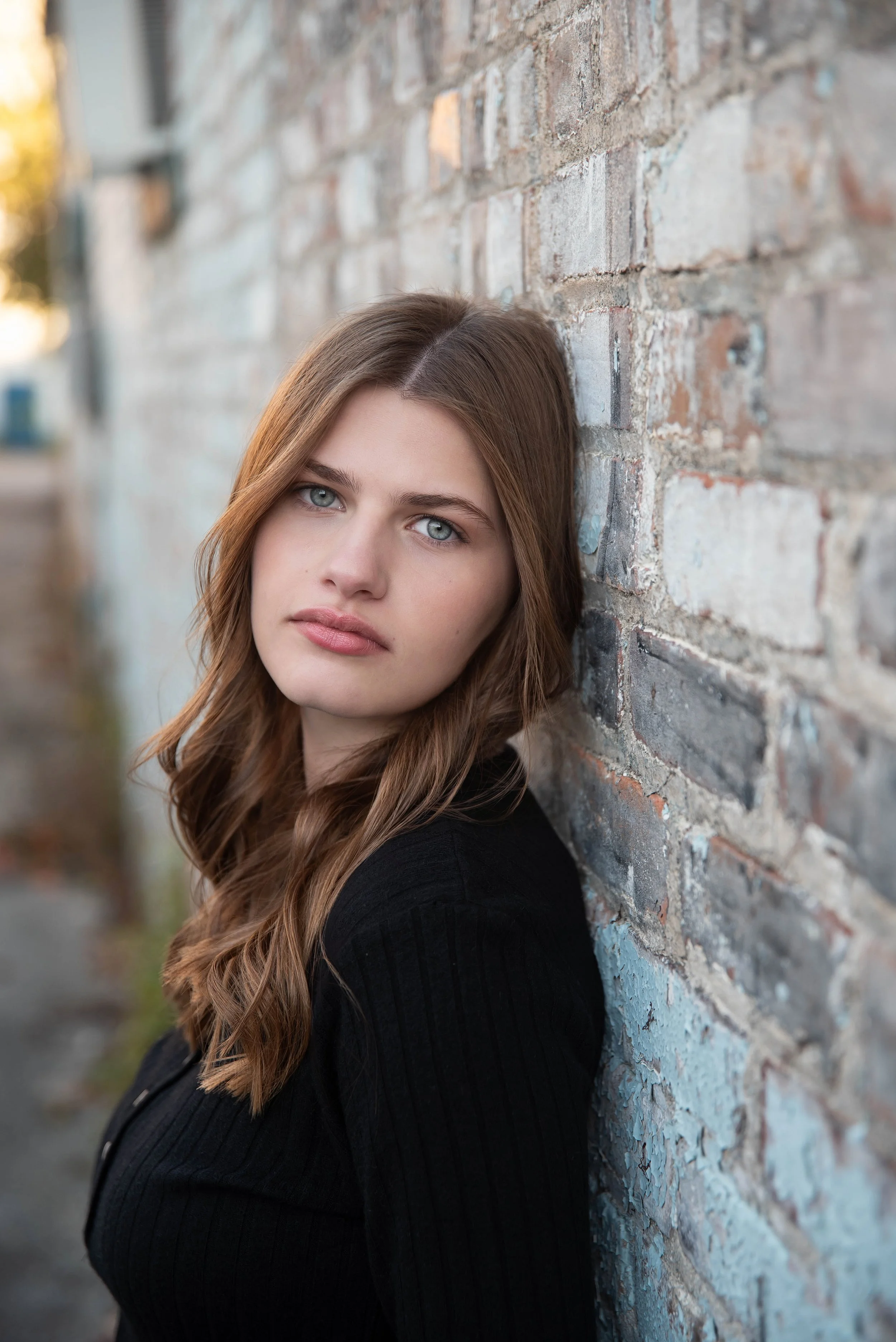 A young woman with long, wavy brown hair and blue eyes leaning against a weathered brick wall outdoors.