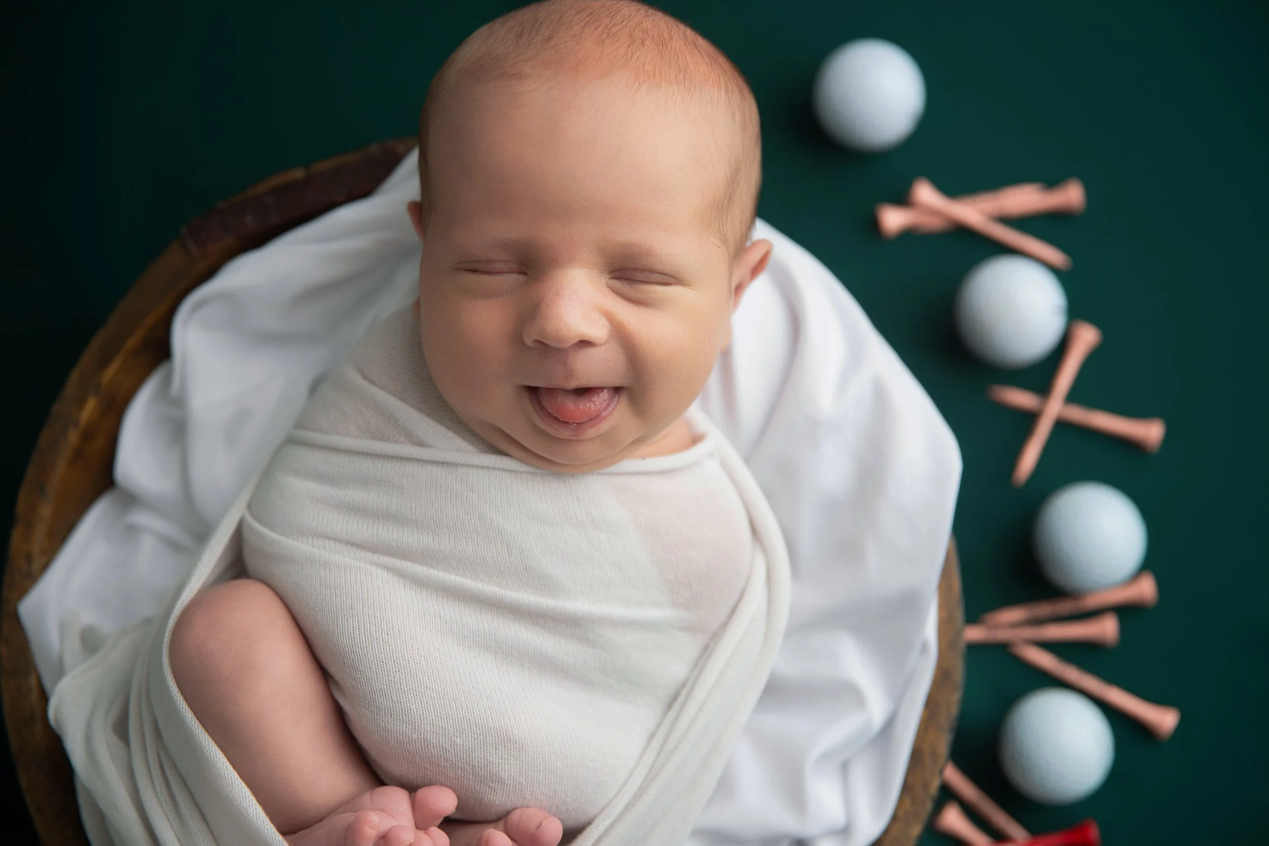 Smiling baby with closed eyes, dressed in white, lying in basket on white cloth with golf balls and sticks in the background.